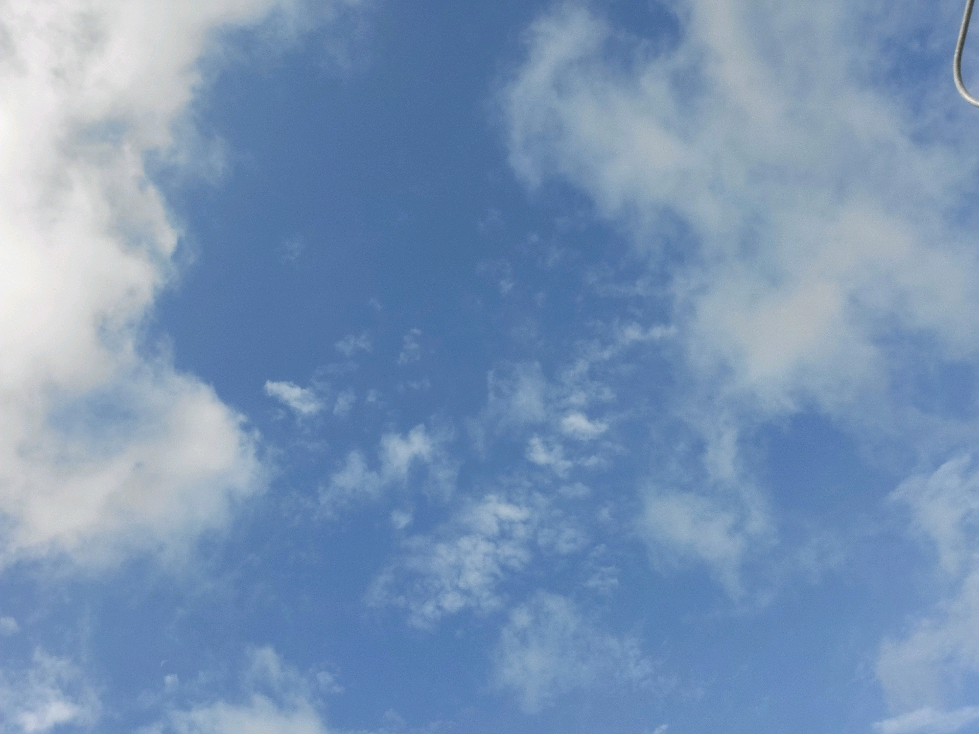 The image depicts a blue sky with scattered white clouds. A thin, curved white object is visible in the upper right corner of the frame. No people, subjects, or specific landmarks of Jelgava, Latvia are discernible within the image. The scene is entirely of the sky.