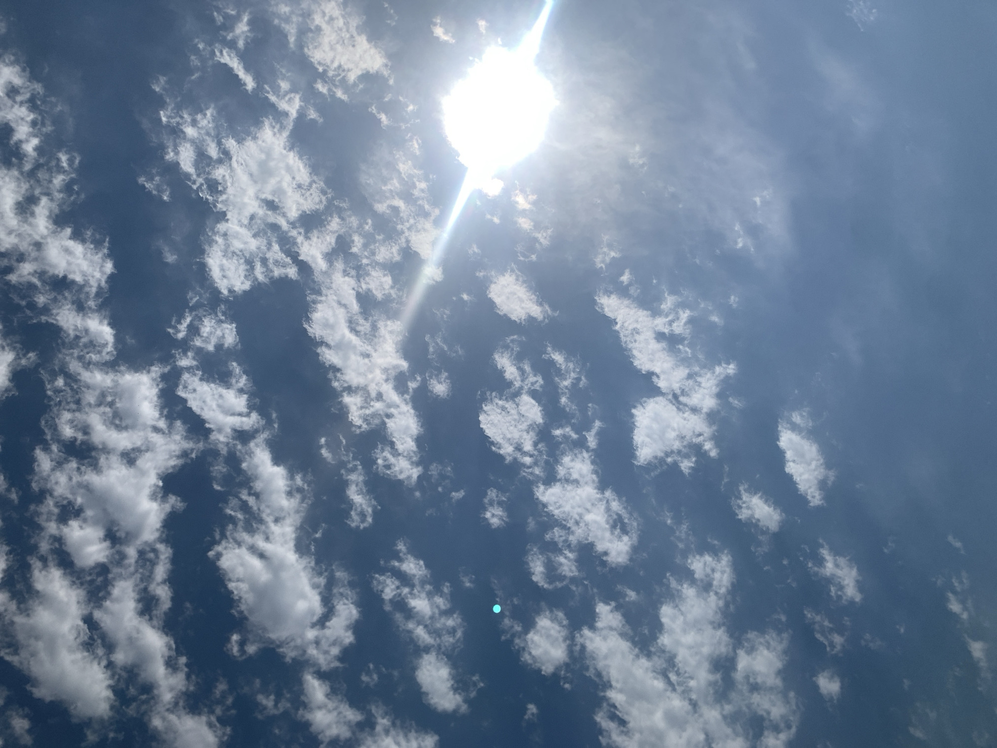 A bright sun is visible in the upper right quadrant of the image, creating a lens flare and a bright beam of light stretching across the sky. The sky is a deep blue, with scattered white clouds. The clouds are wispy and appear to be in rows, creating a textured pattern across the sky. One small, circular, light blue object is visible in the left quadrant of the image. The image appears to be taken from the ground, looking upwards at the sky. The location is Khazar Raion, Azerbaijan.