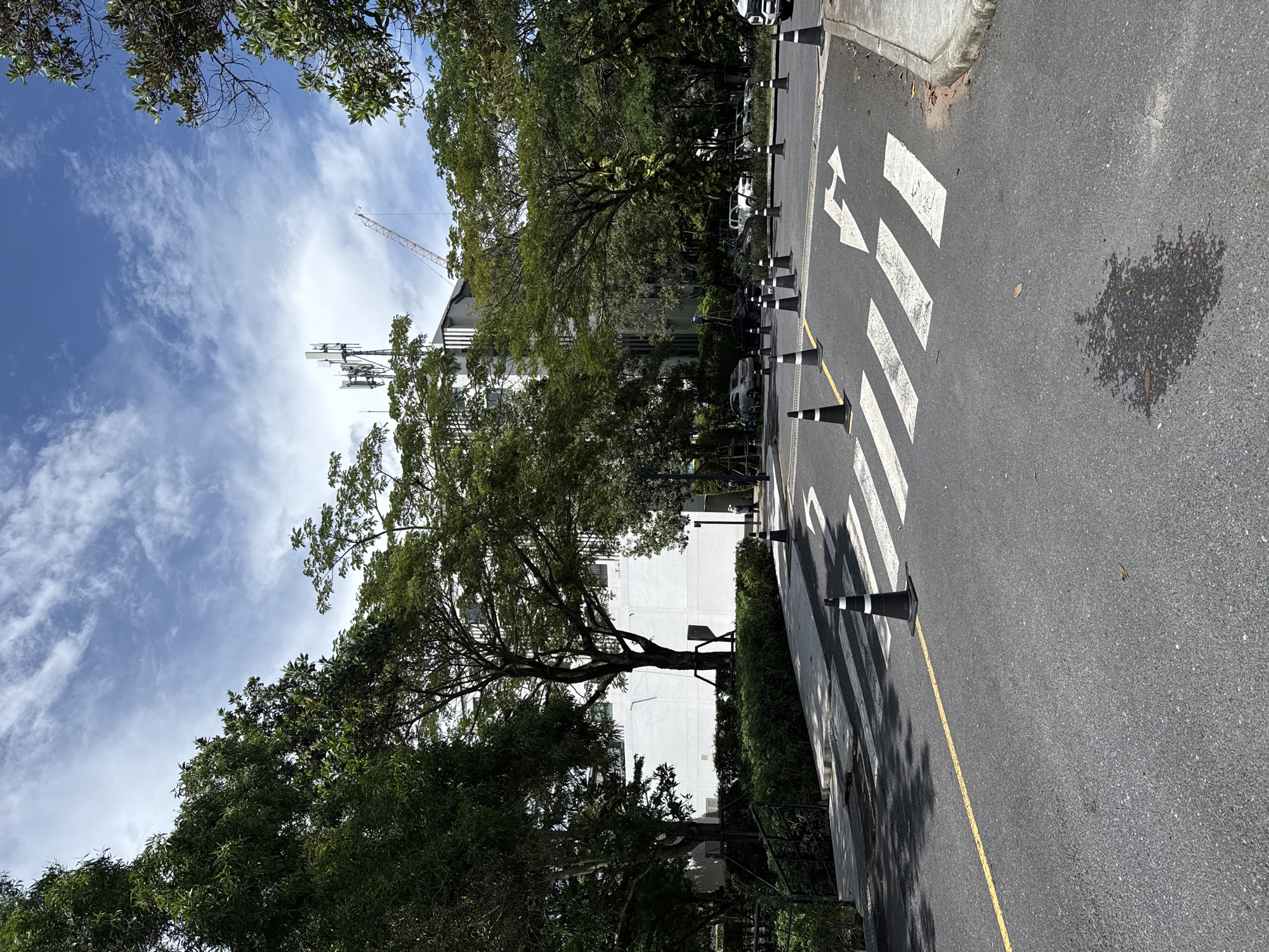 The image depicts a street in Choeng Thale, Thailand, with a crosswalk delineated by white stripes. Black and white striped traffic cones are positioned along the edge of the road. A yellow line marks the lane. Trees line the side of the road, partially obscuring a white building. In the distance, a construction crane and a communications tower are visible against a blue sky with scattered clouds. The asphalt shows signs of wear, including a dark spot.