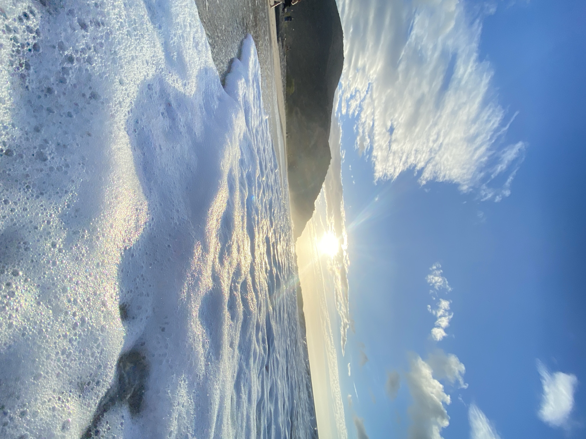 The image depicts a beach scene in Pedro González, Venezuela, on a sunny day. A wave with extensive foam is crashing onto the shore. In the background, there is a mountain. Above, the sky is blue with scattered white clouds. The sun is visible, emitting strong rays. There are people on the beach in the upper-right part of the picture.