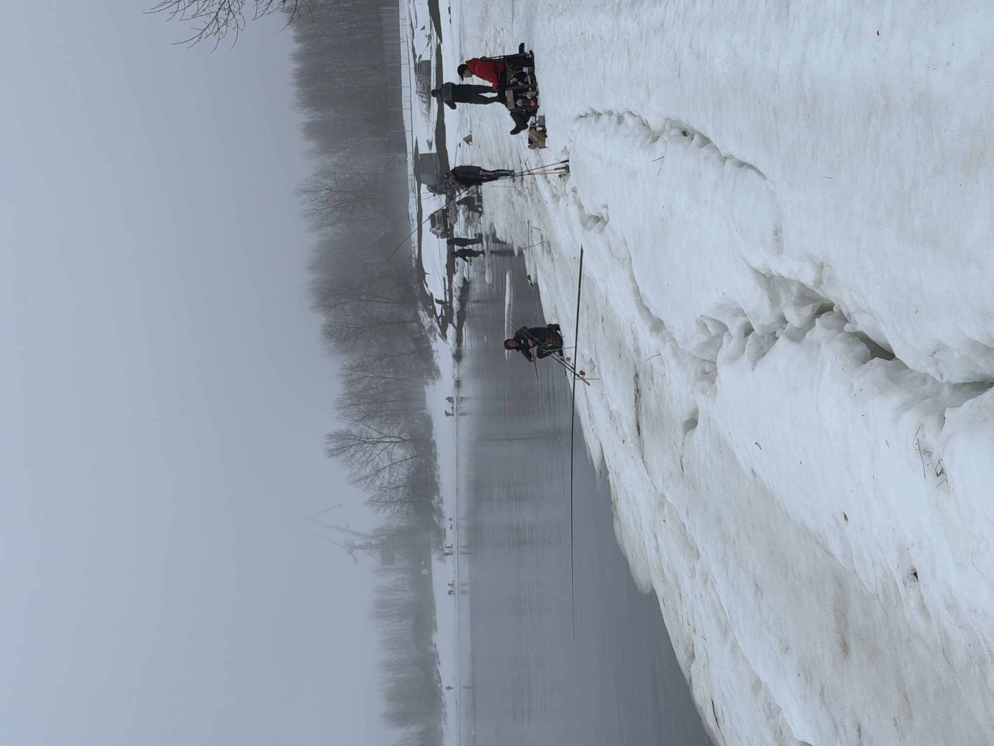 The image depicts a group of people ice fishing on a snowy bank along a body of water on a foggy day in Chaikovsky, Russia. Several individuals are standing near the edge, holding fishing rods. One person is seated in what appears to be a modified wheelchair with equipment. The snow-covered bank is prominent in the foreground. The water extends into the distance, obscured by fog. Bare trees line the opposite shore, with a crane visible through the mist.