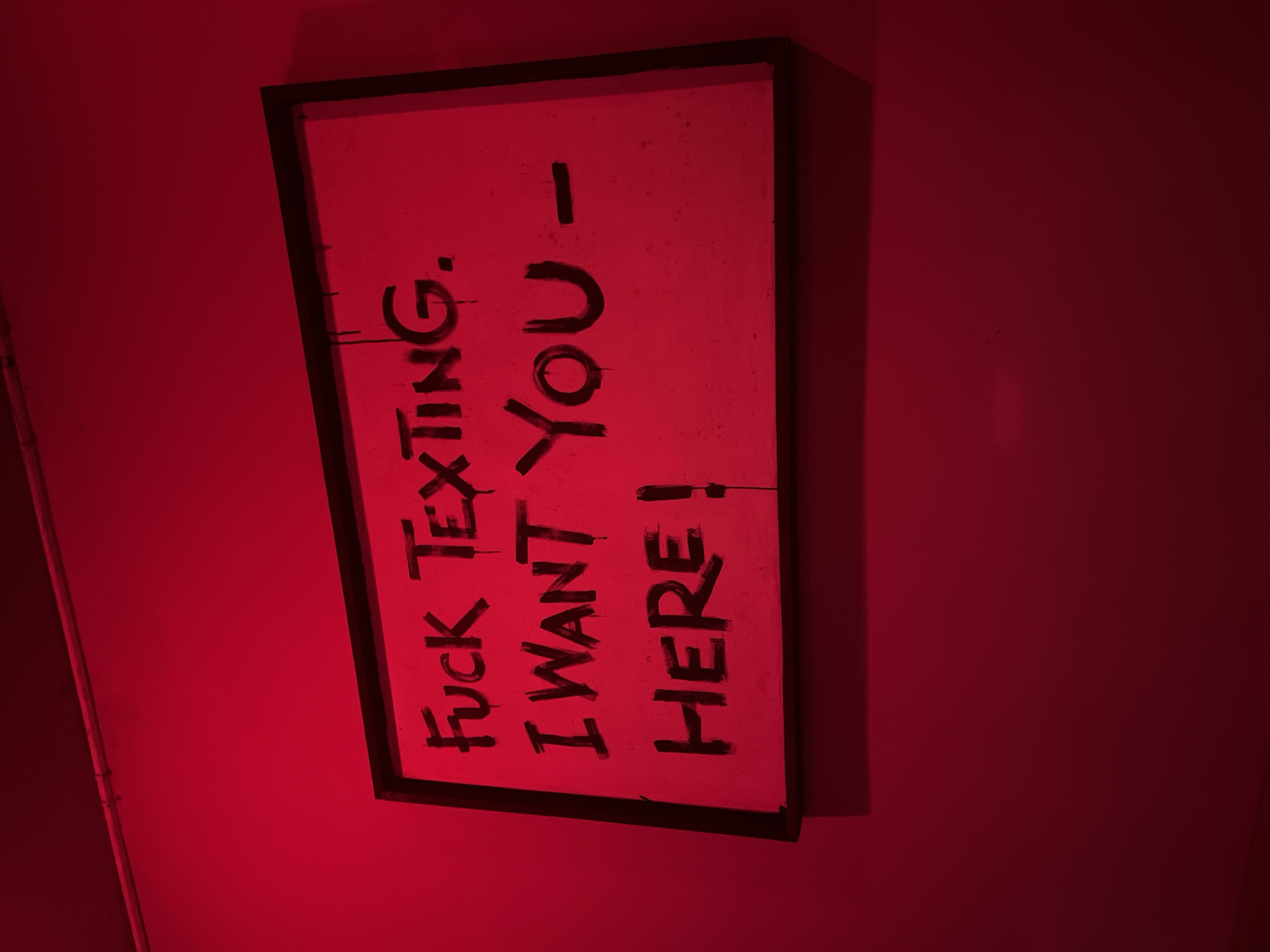The image features a rectangular framed sign, mounted vertically on a wall, bathed in an intense, deep red light. The photograph itself is rotated 90 degrees clockwise, causing the black text on the sign to appear running vertically from the top left to the bottom right of the image frame. The sign's surface, likely white or light-colored, is entirely washed in a strong pinkish-red hue due to the dominant illumination. The background wall is a uniform, dark crimson red, reflecting the dramatic lighting.
The text on the sign is hand-painted in bold, uppercase black letters, exhibiting some visible drips and an uneven quality, which adds to a raw and expressive aesthetic. The full text reads: "FUCK TEXTING. I WANT YOU - HERE!". This direct message conveys a sense of urgency, passion, or longing. The sign is enclosed in a simple black frame that contrasts with the vibrant red backdrop. This scene, located in North Kuta, Indonesia, suggests an urban, artistic, or nightlife environment, possibly a bar or gallery, characterized by its striking color palette and bold statement.