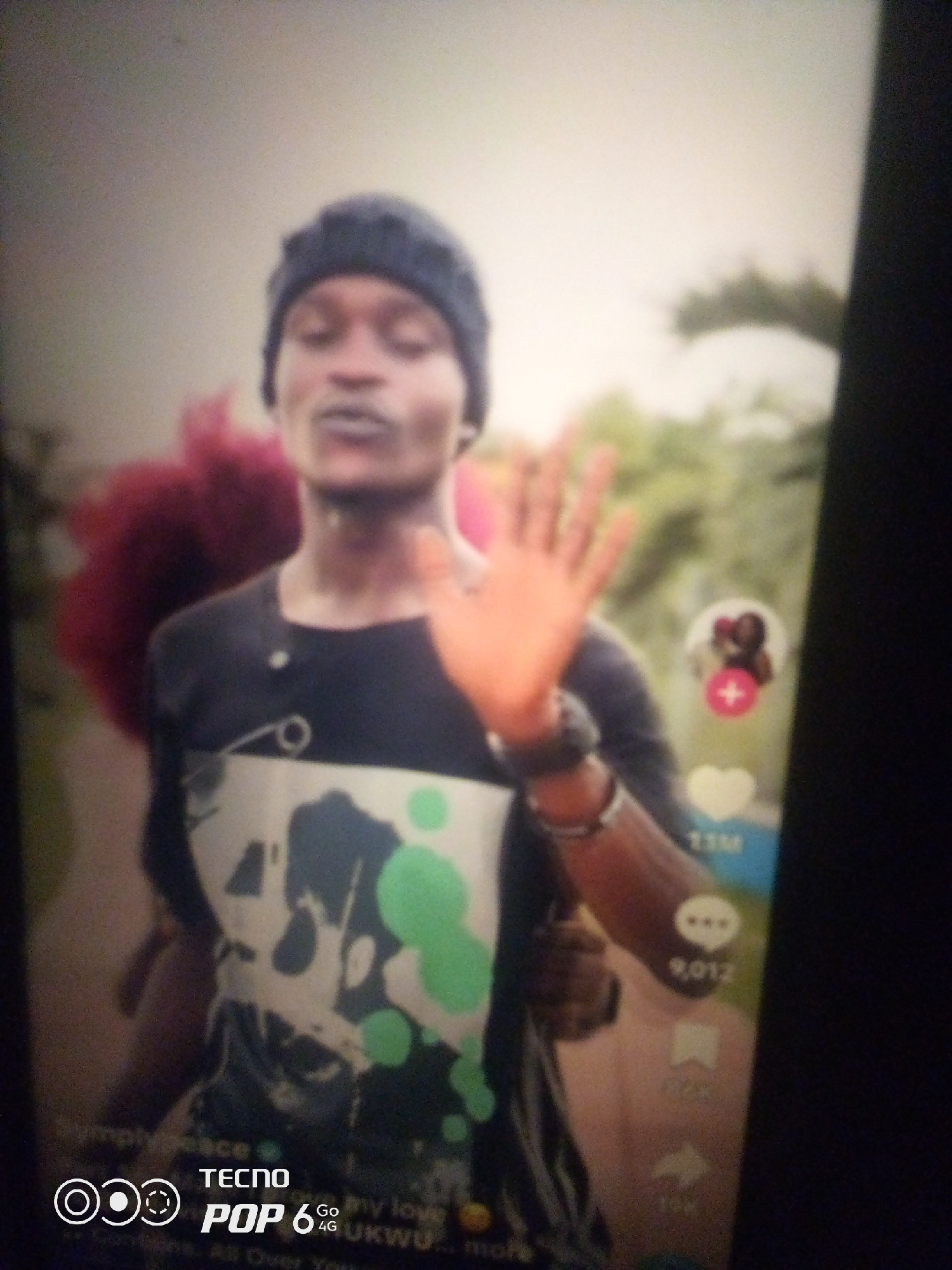 A man wearing a dark beanie and a black t-shirt with a white graphic design is holding up his right hand. He has a short beard and is looking forward with his mouth slightly open. The background appears to be outdoors with greenery. On the right side of the image, there are social media icons, including a heart and a comment icon with a number next to it. The bottom of the image displays "TECNO POP 6 4G UKWU". The scene does not provide any specific details that would indicate its location within Howayi, Nigeria.