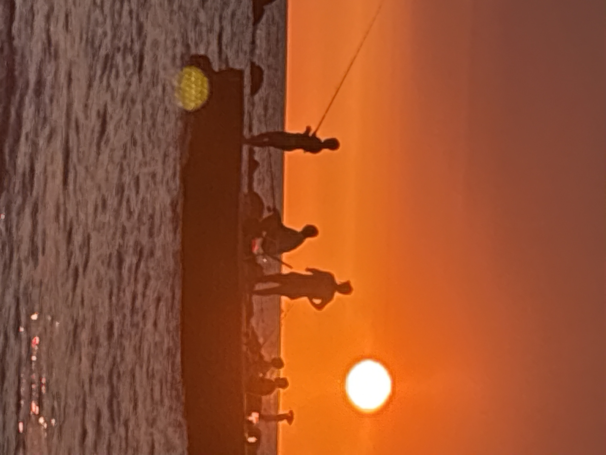 A group of people are silhouetted against an orange sunset, fishing from a pier in the city of Bar, Montenegro. The sun, a bright white disk, is positioned high in the sky to the right of the frame. Several individuals, some holding fishing rods, are scattered across the pier. The water, rippled and dark, reflects the warm hues of the sunset. A yellow, textured object, possibly a buoy, floats in the water to the left of the pier. The sky transitions from a deep orange at the horizon to a reddish-brown at the top of the image.