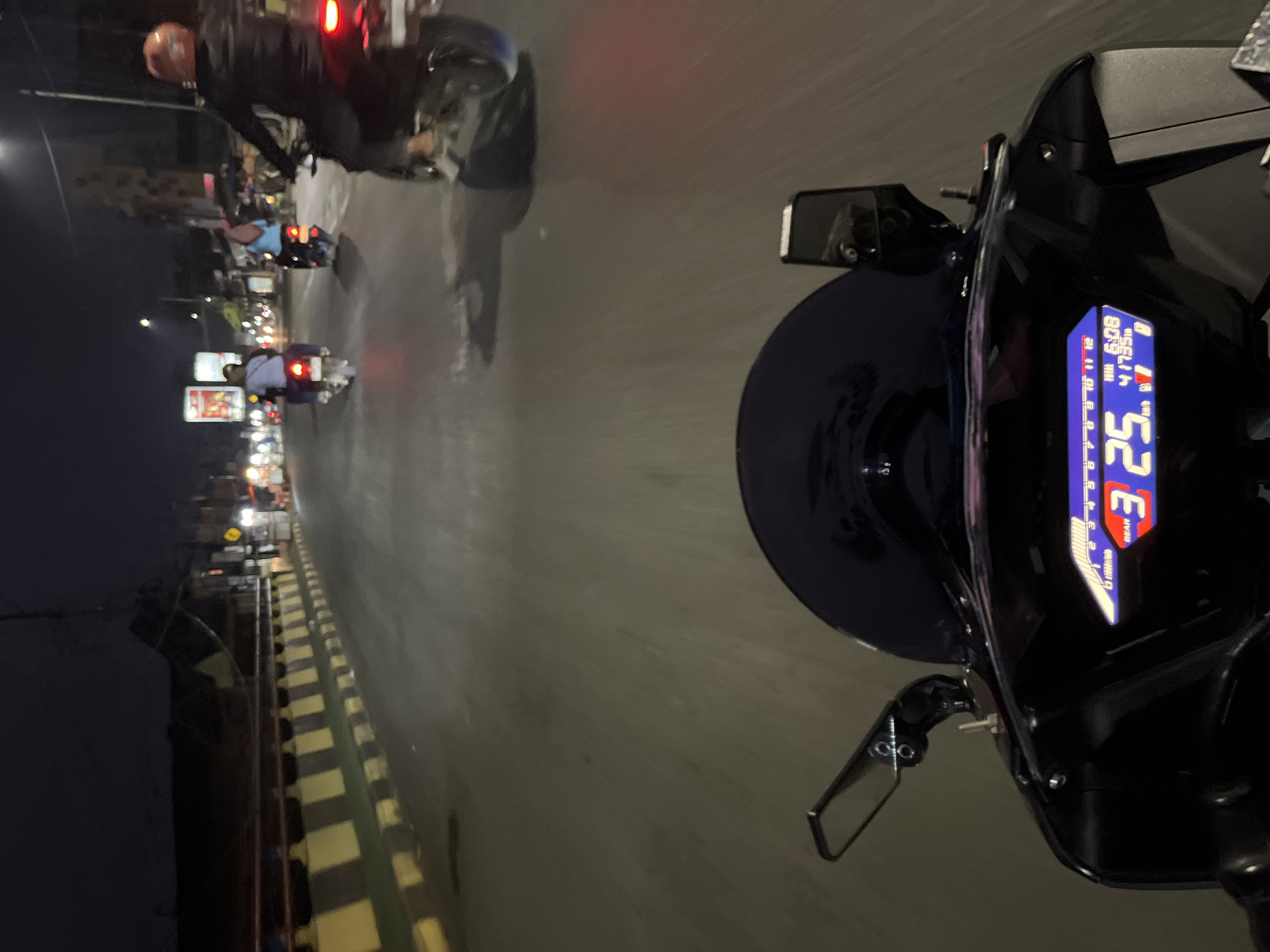 This is a first-person perspective, taken from a motorcycle at night, on a paved road, likely in Pecangaan, Indonesia. The road is lined with a yellow and black striped barrier on the left, and buildings are visible in the background. Several other motorcycles are on the road, with riders wearing helmets. One motorcycle in the foreground has a digital display showing "25" for speed, "3" for gear, "6:08" for time, and "41735" for mileage. The lighting suggests streetlights and the headlights/taillights of vehicles. The overall atmosphere is that of nighttime traffic in an urban or semi-urban area.