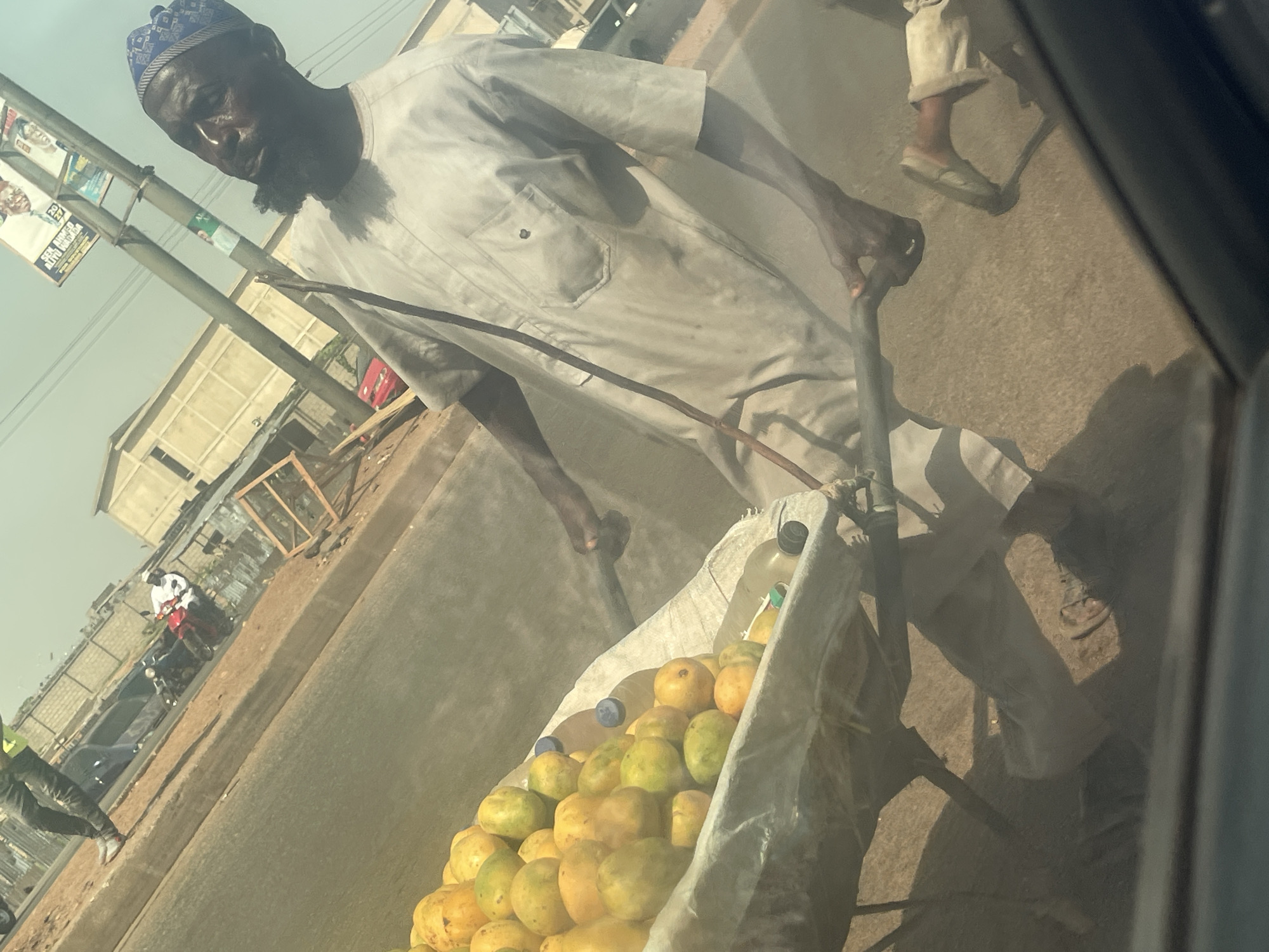 This image depicts a vibrant street scene in Keffi, Nigeria, likely during the daytime under dry, possibly hazy conditions. The main subject is a dark-skinned man, appearing to be in his 40s or 50s, with a short beard. He wears a light-colored, long-sleeved tunic, typical of Nigerian traditional attire, and a blue and white patterned cap. He looks directly towards the camera with a neutral expression.
The man is actively pushing a metal wheelbarrow or handcart, which is laden with numerous yellow and green fruits, likely mangoes or oranges, nestled within a white fabric lining. Two plastic bottles, one with a blue cap and one with a black cap, containing clear liquid, are also visible among the fruit. This suggests he is a street vendor.
The scene takes place on a road, which appears to be paved or hardened earth, with a visible curb. In the background, low-rise buildings, some constructed with concrete blocks, line the street. Utility poles stand along the roadside; one pole displays several political posters. Visible text on these posters includes "ALL Progressives Congress" (APC), a major political party in Nigeria, and phrases such as "AGAIN FOR THE PEOPLE," indicating a political campaign.
Other people are present in the environment: a person in a bright yellow-green vest and dark pants is partially visible walking on the far left, and another person can be seen on a red motorcycle or scooter further in the background. The photograph appears to have been taken from inside a vehicle, evidenced by the dark frame on the right side. The overall lighting is warm, contributing to the sense of a busy, sunny, or dusty day characteristic of the region.