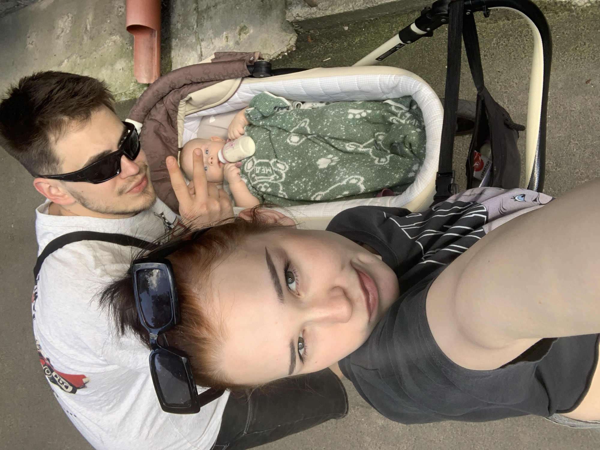 The image shows a young couple with a baby in a stroller, likely taken in Kyiv, Ukraine. The man, wearing sunglasses and a white t-shirt, is holding a baby bottle to the baby's mouth. The baby, covered with a green blanket with a paw print pattern, is lying in the stroller. The woman, in a black t-shirt, is lying next to the stroller, looking at the camera. The background shows a concrete surface and a reddish pipe, suggesting an outdoor urban setting. The lighting indicates it is daytime. The overall mood appears casual and familial. There is no visible text in the image.