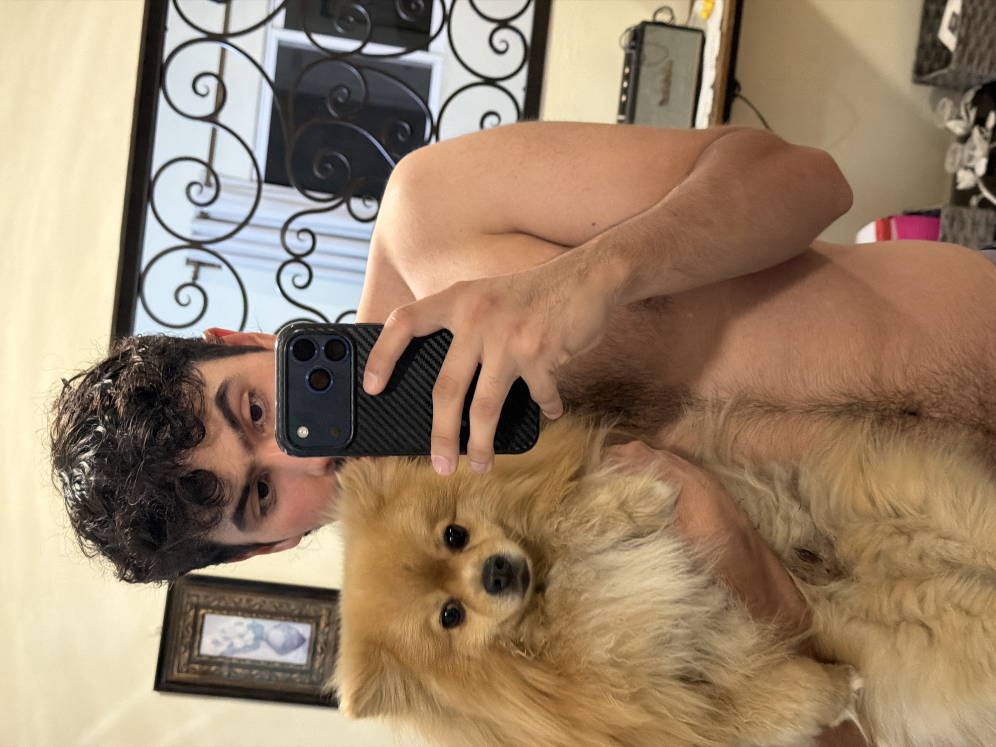 A shirtless man with dark, curly hair holds a fluffy, golden-brown Pomeranian dog while taking a selfie in front of a mirror. The man is holding a black iPhone with a carbon fiber case. The background includes a wrought-iron mirror frame, a framed picture on the wall, and a glimpse of other furniture and decor. There is no evidence to suggest the location is Cuautla, Mexico.