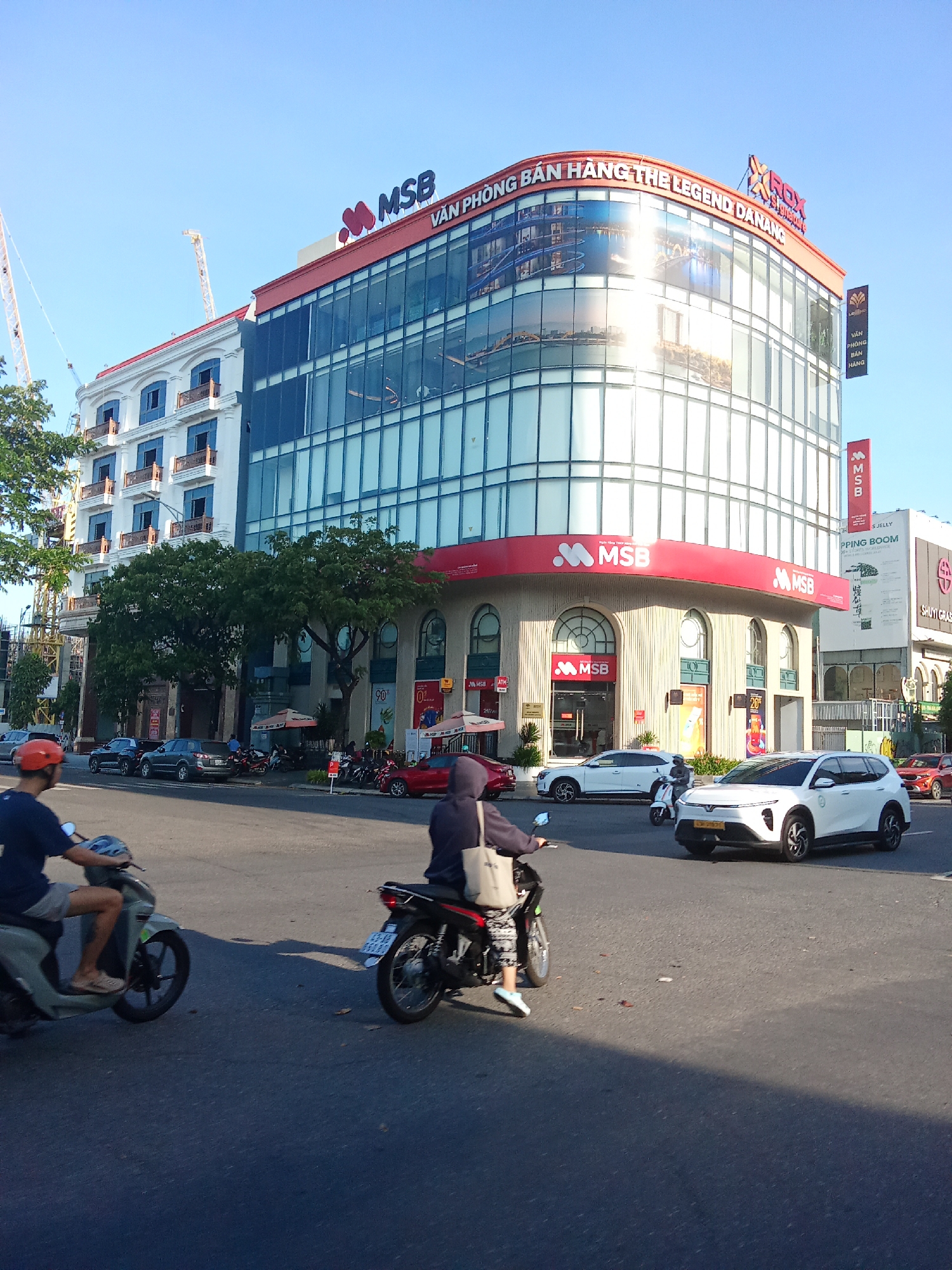 The image captures a street scene in Da Nang, Vietnam, featuring a modern building with prominent signage. The building has a curved facade with large glass windows and a red accent band. On top, the words "MSB" are visible in large blue letters, along with a red heart logo. Above this, the phrase "VĂN PHÒNG BÁN HÀNG THE LEGEND ĐÀ NẴNG" is displayed, indicating it's a sales office. A smaller "ROX" logo is also present.
Several vehicles are on the street, including motorcycles and cars. Two individuals are riding motorcycles in the foreground. One rider, wearing a blue shirt and shorts, is on a light-colored scooter. The other rider, in a dark hoodie and patterned pants, is on a black motorcycle. White cars are also visible, including one with a license plate that appears to be a yellow tourist plate.
The environment suggests a sunny day with a clear blue sky. Trees line the street, and construction cranes are visible in the background, suggesting ongoing development. The building also displays a vertical red banner with "MSB" on it. There are awnings over some ground-level entrances, and umbrellas are set up outside some establishments. The overall impression is a bustling urban environment with commercial activity.