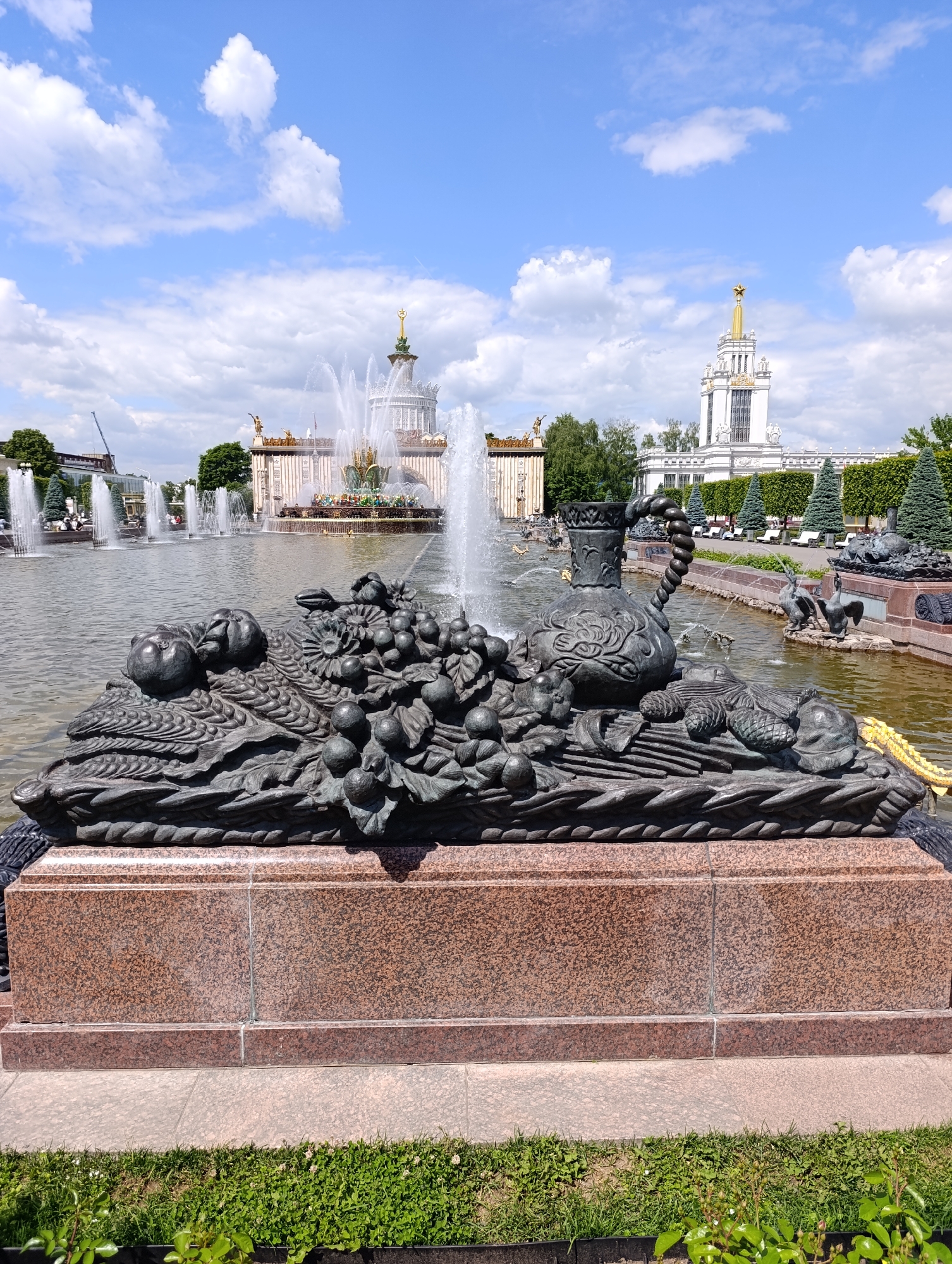 This image captures a vibrant outdoor scene at the Exhibition of Achievements of National Economy (VDNKh) in Moscow, Russia, on a bright, sunny day with a clear blue sky dotted with white clouds.
In the immediate foreground, a large, dark bronze sculpture of a decorative basket or cornucopia overflows with various fruits and produce, including grapes, apples, and what appear to be pineapples or other exotic fruits, alongside a stylized urn or jug. This sculpture rests on a sturdy base made of reddish-brown speckled granite. Beneath it, a strip of lush green grass with small white flowers is visible.
The mid-ground features a wide body of water, likely a large pond or canal, from which numerous fountains powerfully spray water high into the air. Smaller, intricate sculptures, including figures resembling geese, are also visible within the water or along its edges.
In the background, several grand and ornate buildings with classical and Soviet-era architectural styles dominate the skyline. The most prominent building on the left features multiple spires and domes, with a golden spire topped by a star. Another large, white building with a similar golden star-topped spire stands further to the right. Both structures are characteristic of Stalinist Empire style architecture found at VDNKh. Lush green trees, carefully trimmed hedges, and manicured landscaping surround the buildings and line the pathways. Small figures of people can be seen in the distance, strolling and visiting the complex, indicating a bustling public space.
The overall scene depicts a lively and picturesque park or exhibition ground, with the active fountains and decorative elements creating a sense of grandeur and historical significance. No discernible text is visible in the image.