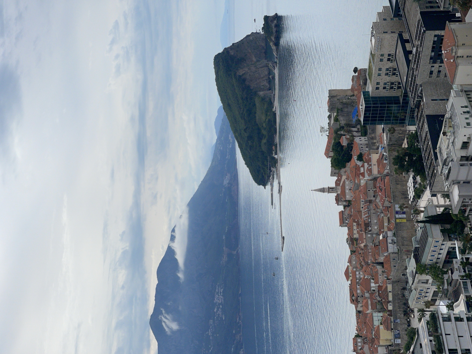 The image depicts the coastal cityscape of Budva, Montenegro. In the foreground is the old town with its red-tiled roofs and the modern architecture near the shoreline. The Adriatic Sea separates the town from Sveti Stefan island, which is visible in the distance. Further inland, a mountainous landscape extends into the background with one peak partially obscured by clouds. The sky is overcast.