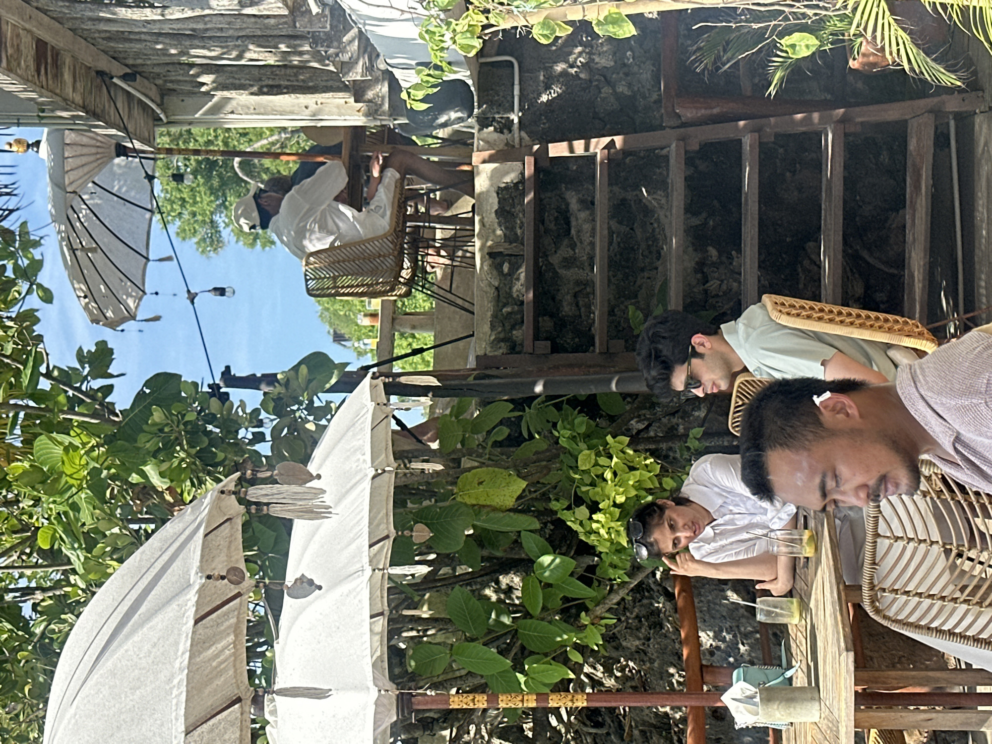 The image shows a group of people sitting at an outdoor cafe in Kuta Selatan, Indonesia. The cafe has a rustic, tropical vibe, with white umbrellas providing shade. Three people are seated at a wooden table, and their conversation is light. One is in a light purple shirt in front of the table, another is wearing a light green shirt, and another is wearing a white shirt. The cafe is built into a rocky hillside. In the background, a man is sitting under a similar umbrella. The setting appears to be a relaxed, tourist-friendly location.