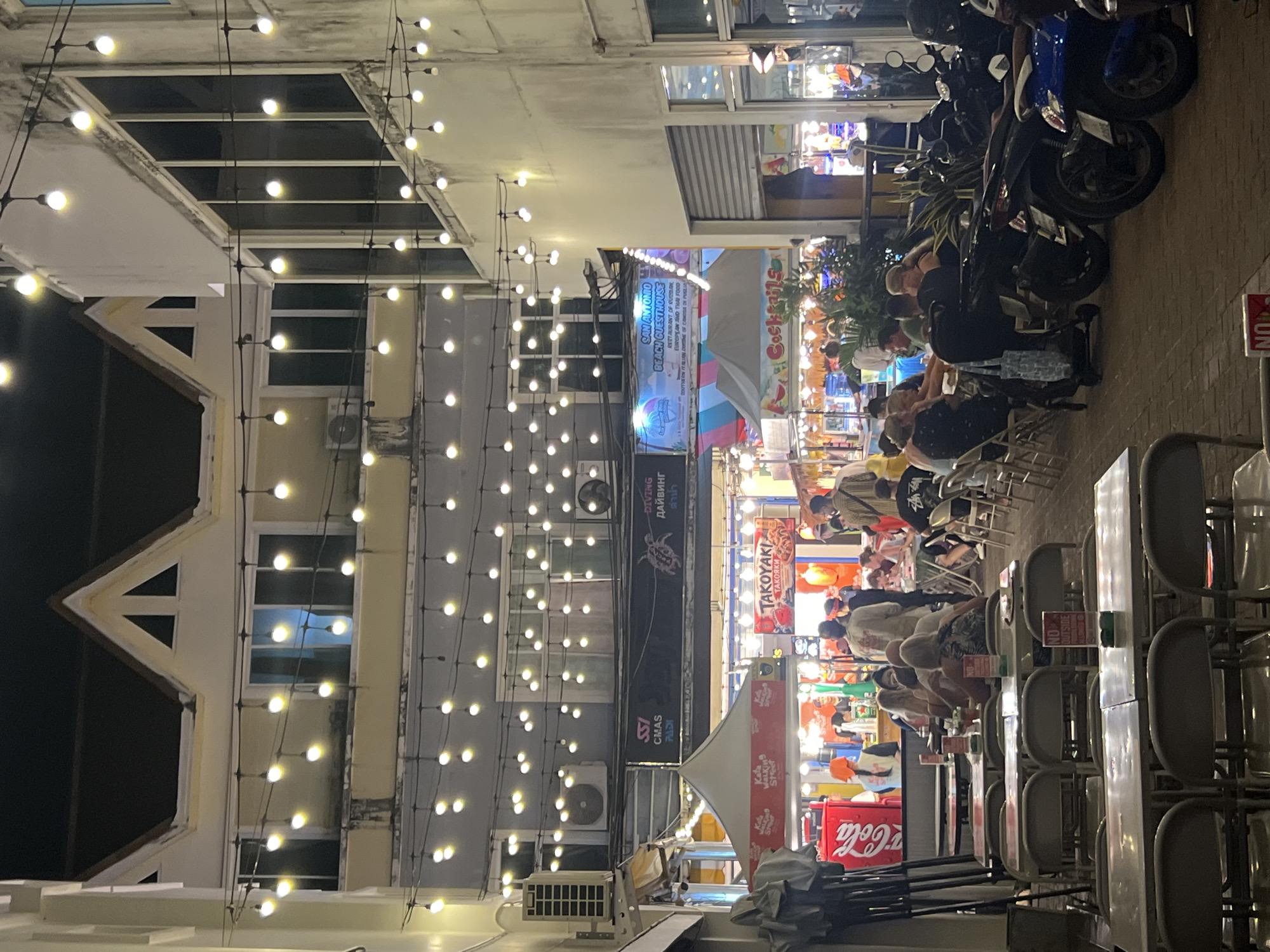 This image depicts an outdoor scene at night, likely a street market or a restaurant area in Karon, Thailand. String lights illuminate the area, casting a warm glow over several buildings and outdoor seating arrangements. Several people are visible, mostly seated at tables, suggesting they are dining or socializing. In the background, motorcycles are parked, indicating a mode of transportation common in the area.
The scene is bustling with activity, with various stalls and shops visible, some displaying signs with text. Notable text includes "Cola," "ISS CMAS PADI," "Kata Street," "TAKOYAKI," "DIVING," "SAN ANTONIO BEACH GUESTHOUSE," and descriptions of food offerings. The presence of air conditioning units on the building walls suggests a tropical climate. The overall atmosphere appears lively and casual, typical of a tourist-oriented evening setting. The lighting and the presence of people enjoying themselves suggest a pleasant evening atmosphere, possibly warm and humid given the tropical location.