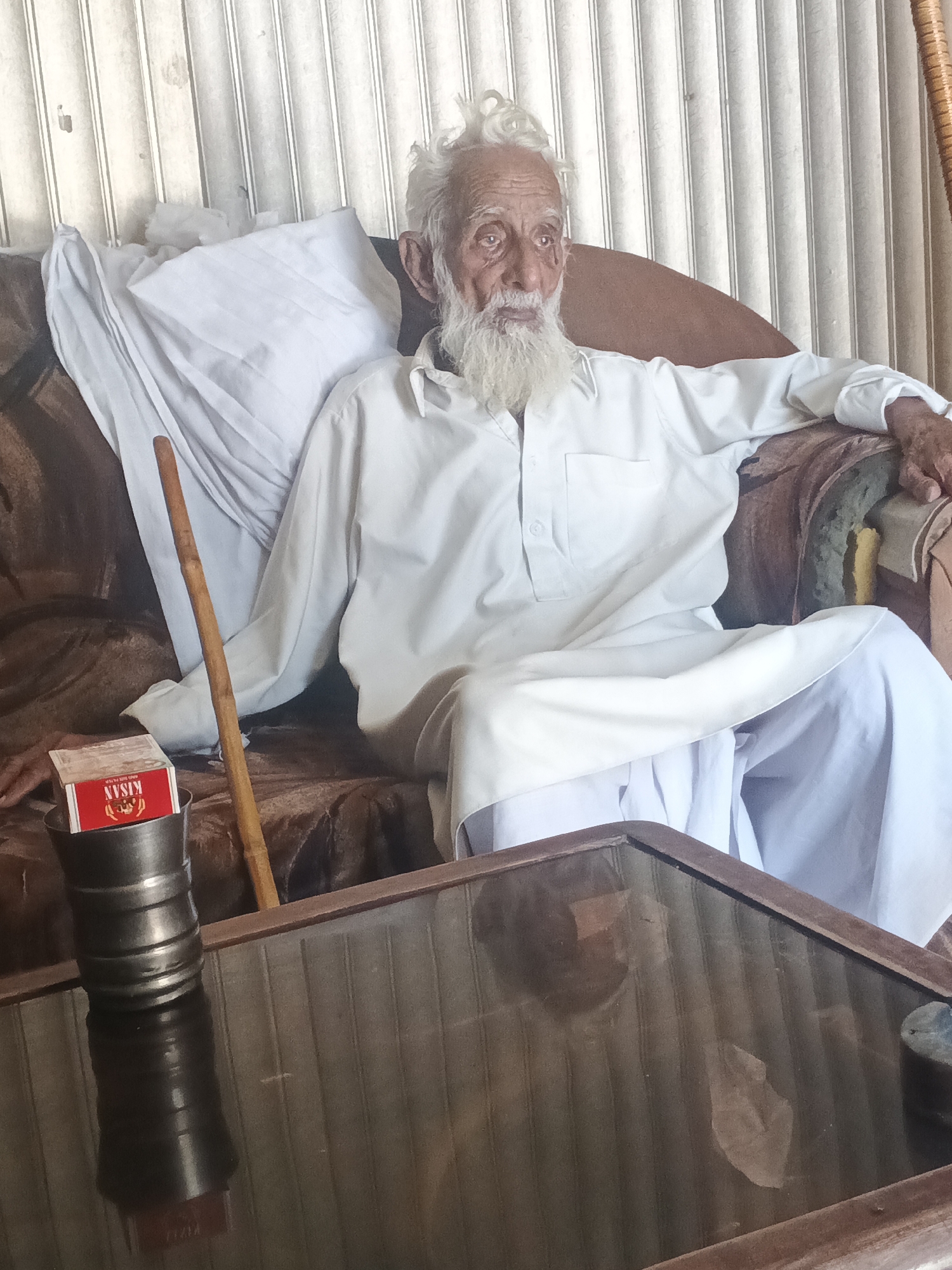 An elderly man with white hair and a long white beard sits on a brown couch indoors, likely in a domestic setting. He is dressed in white clothing. A wooden walking stick leans against the couch next to him. A metal cup with a red and white "Kisan" brand box sits on a small table in front of him, reflecting on the glass surface. Vertical blinds are visible on the wall behind the couch. The location is Samundri, Pakistan.