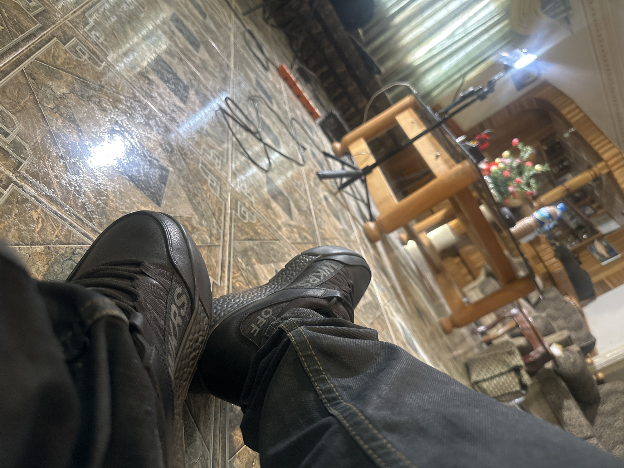A first-person view of the legs of a person wearing dark jeans and black sneakers sitting indoors. The sneakers have "OFF" and "WKRS" printed on them. The floor is tiled in a brown and beige pattern. A wooden loom sits in the background with a lamp attached. Cables run along the floor. Through an archway in the background are blurry interior details, including a Christmas tree. The location, Jos, Nigeria, is not discernible from the image.