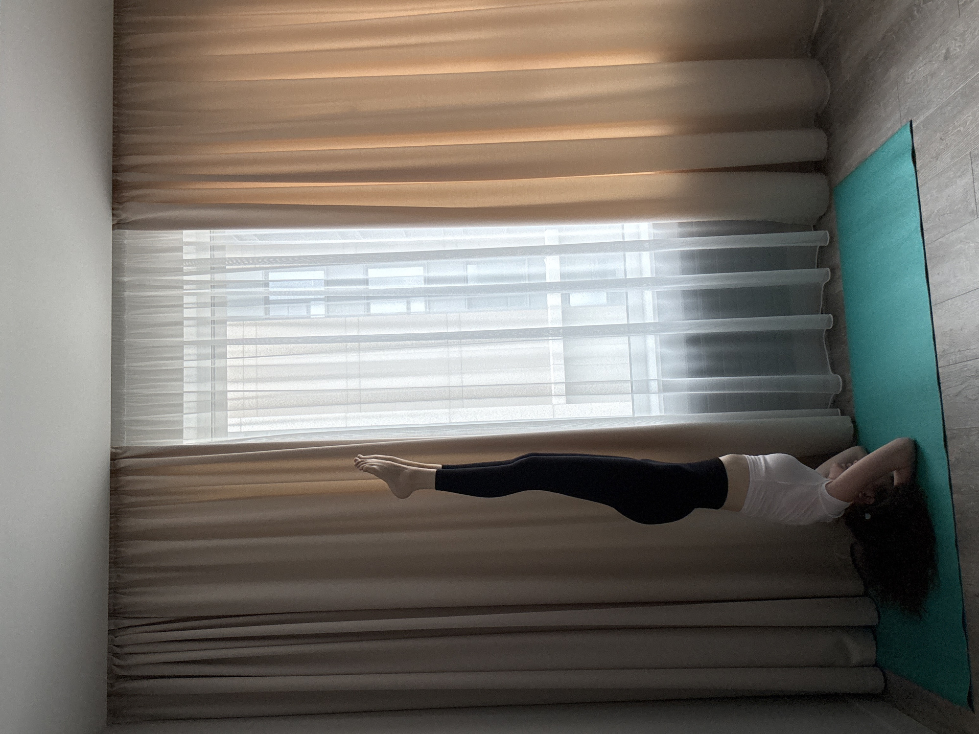 A person is performing a headstand on a turquoise yoga mat on a wooden floor. The person is wearing a white t-shirt, white shorts, and black leggings. Their legs are extended upwards, and their arms are bent, supporting their head on the mat. Their hair is spread out around their head. The scene is indoors, with large beige curtains covering a wall on the left and right, and sheer white curtains in the center covering a window. The lighting suggests it is daytime. There is no indication of the location in Moscow, Russia.