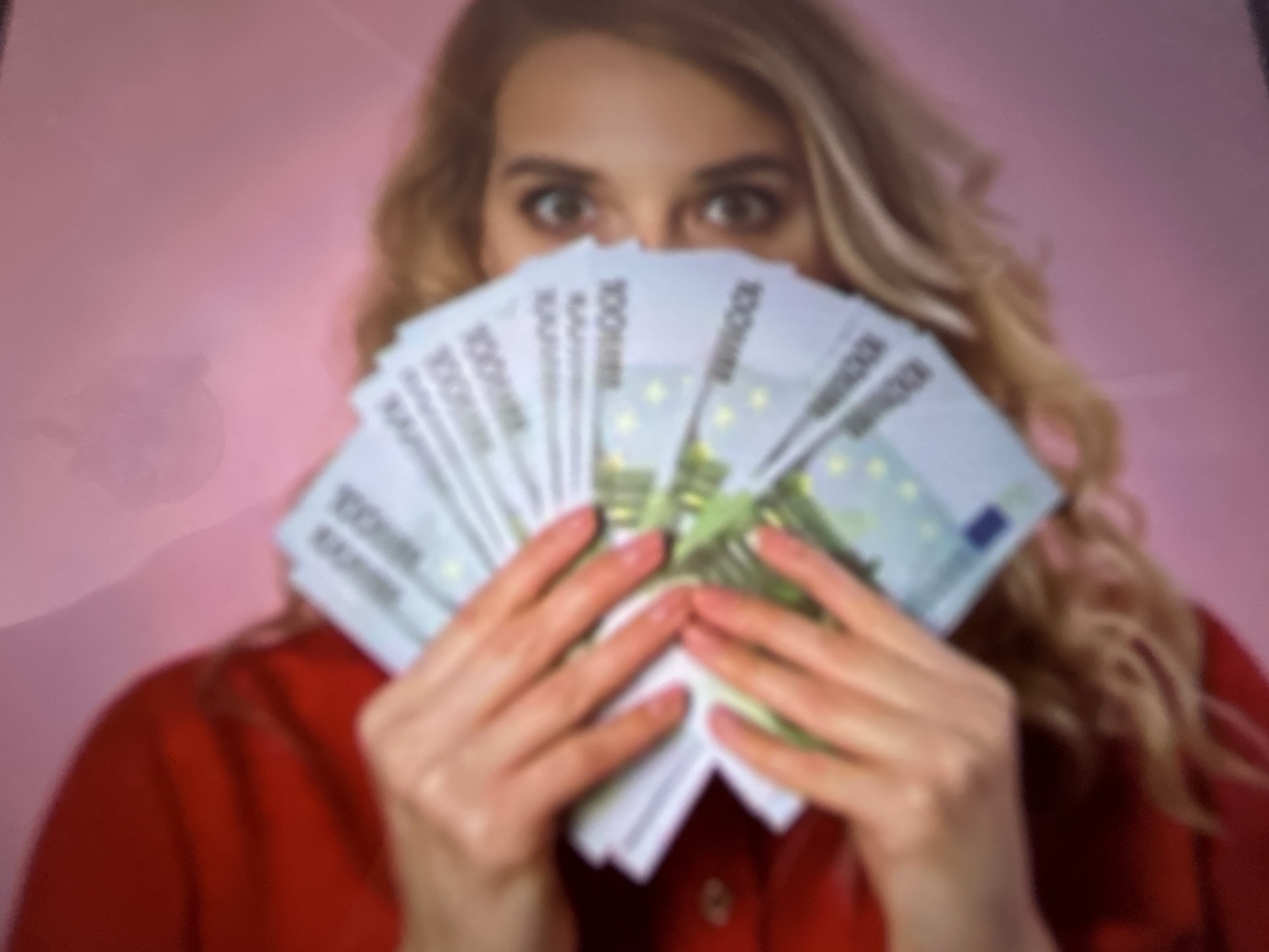 The image shows a person holding a fan of 100 euro banknotes in front of their face, with only their eyes and a portion of their nose visible. The person has blonde hair and is wearing a red garment. The background is a soft pink color. The image is blurry and appears to be taken from a slightly elevated angle. There is no indication of the city of Gelendzhik, Russia, in the image.