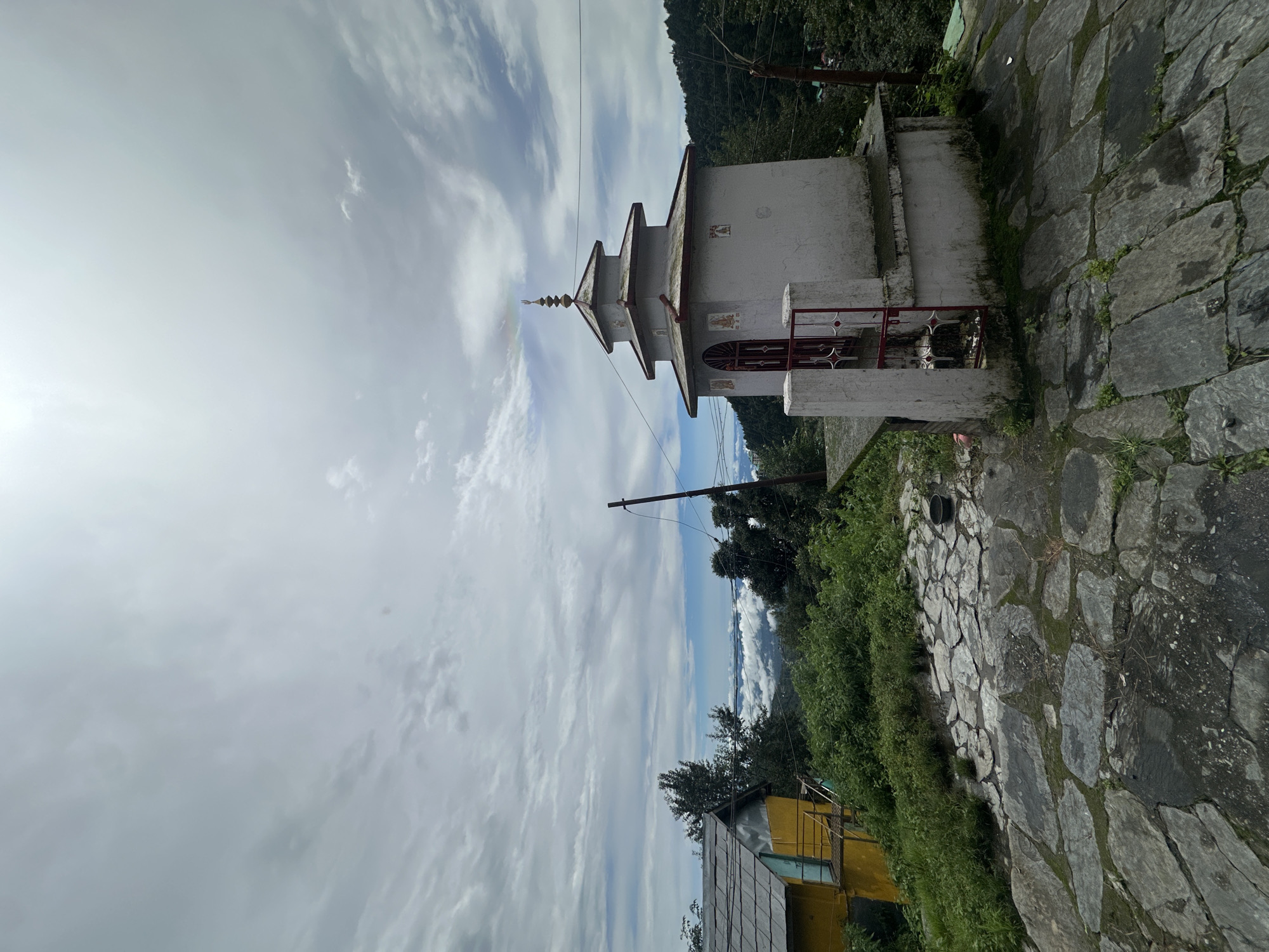 In Haini, India, a small, multi-tiered temple stands prominently on a stone-paved platform. The temple is white with a red-accented door and roof trim, topped with a spire. To the left of the temple, a yellow building with a grey tiled roof is visible, partially obscured by lush green foliage. The background reveals a dramatic vista of rolling hills covered in dense forest, with a layer of white clouds blanketing the valleys below, indicating a high-altitude location. The sky above is overcast with textured clouds. A wooden utility pole with wires stands to the right of the temple. The stone paving in the foreground is uneven, with patches of moss and dampness.