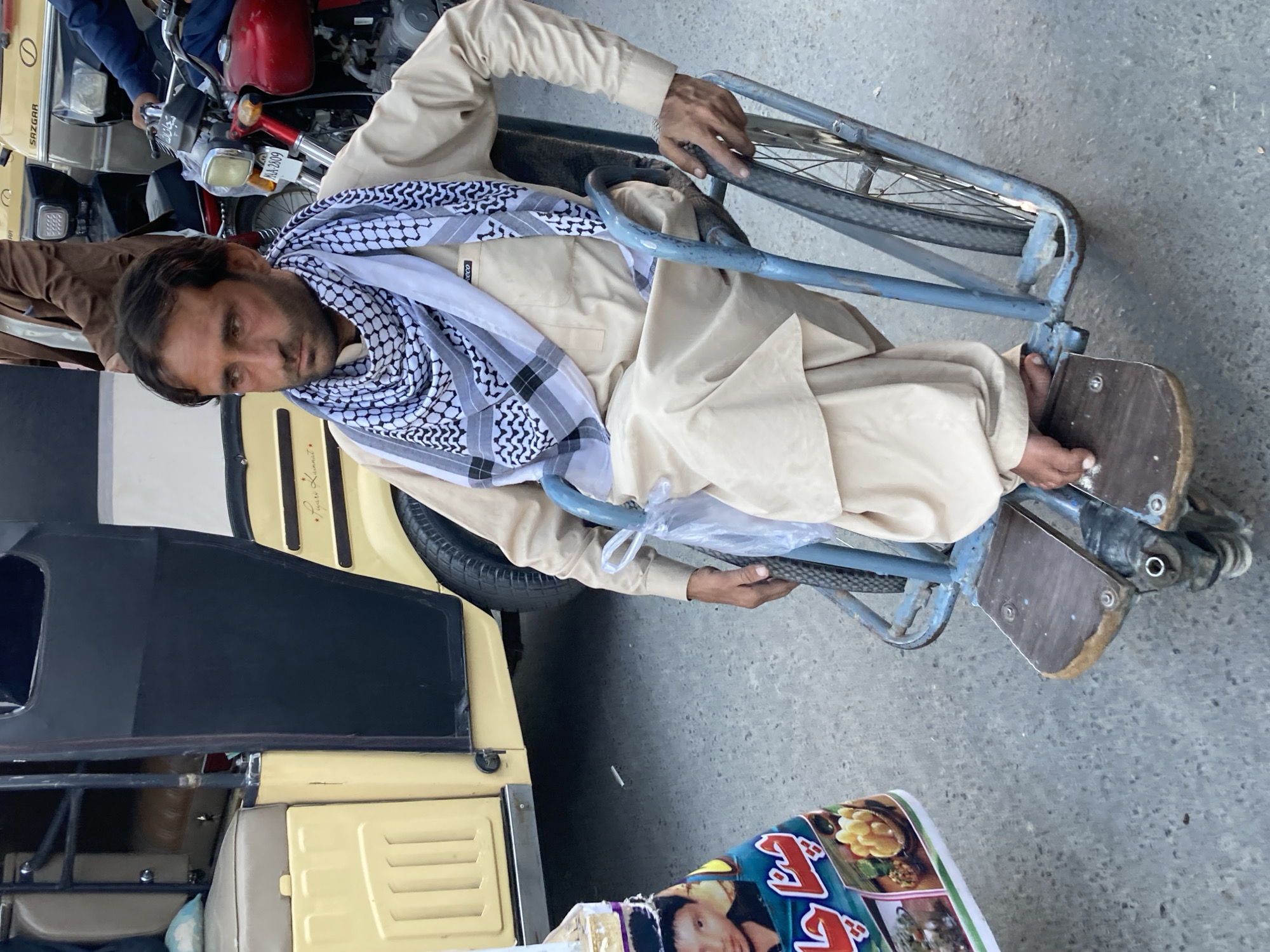 A man wearing traditional Pakistani clothing, including a shemagh scarf, is sitting in a modified wheelchair attached to an auto rickshaw in Quetta City Tehsil, Pakistan. The auto rickshaw is yellow. A motorcycle is nearby. A poster is visible on the ground. The ground appears to be a paved street.