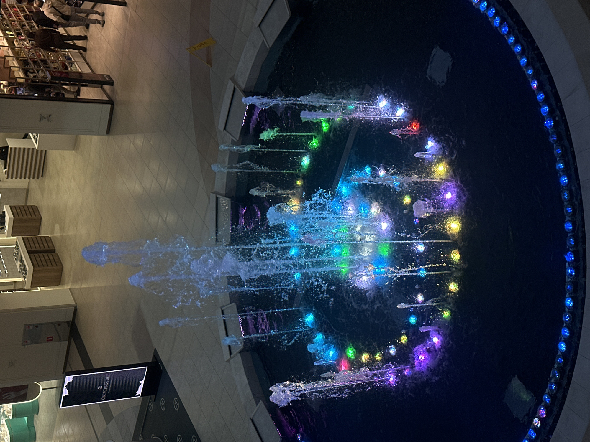 The image displays an indoor scene, likely a shopping mall or a large public atrium in Sochi, Russia, captured from an elevated perspective looking down. The central and most prominent feature is a large, circular or oval-shaped fountain filled with dark water, illuminated by a dynamic and colorful array of LED lights.
Multiple water jets spray upwards and outwards from the fountain, creating various patterns, with the water itself catching the reflections of the vibrant lights. The embedded lights within the fountain glow in shades of blue, green, purple, yellow, and red, providing a lively visual spectacle. The rim of the fountain is lined with a continuous string of bright blue lights, further enhancing its outline.
Surrounding the fountain is a light-colored tiled floor, which extends towards various commercial establishments and shops. Several people are visible in the background, appearing to walk, browse, or stand near shop displays, suggesting a typical public activity. Some wooden benches or seating areas are also present near the shops.
The lighting in the scene is predominantly artificial, indicating that the photograph was taken in the evening or at night, allowing the fountain's light display to be a focal point. A yellow triangular sign, likely a "wet floor" warning, is faintly visible on the tiled floor. A dark hanging sign with white text near the bottom left reads "СИСТЕМА УМНЫХ...", suggesting a "Smart System" in Russian. The overall ambiance is vibrant and engaging.