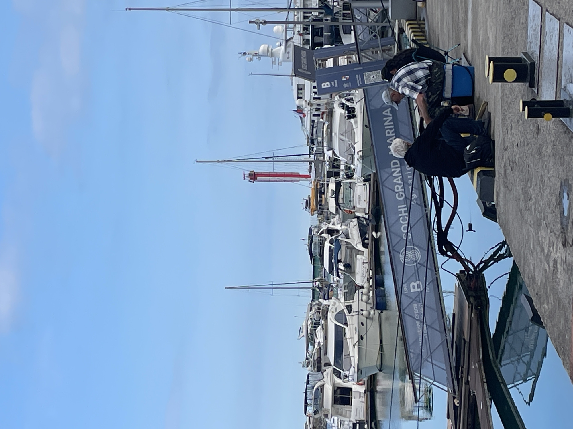 The image depicts a scene at the Sochi Grand Marina in Sochi, Russia, under a clear blue sky with scattered clouds. In the foreground, two men are seated on a concrete pier, engaged in conversation or activity. One man, with gray hair, is wearing a black hoodie and jeans, and is kneeling. The other man is wearing a plaid shirt, a cap, and camouflage-patterned shorts, and is seated on a blue cooler. Beside them are backpacks and fishing gear, suggesting they might be fishing or relaxing.
The pier extends into the water, where numerous yachts and boats are docked. The boats range in size, from smaller leisure craft to larger yachts, many with white hulls and dark-colored tops. The water is calm, reflecting the boats and the sky.
Further in the background, a distinctive red and white tower, possibly a lighthouse or observation tower, is visible among the docked vessels. The marina appears to be a well-maintained facility for sailing and yachting. The overall atmosphere is one of a peaceful day at the harbor.