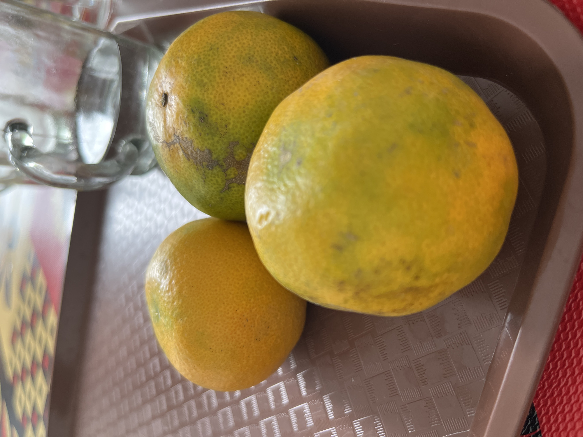 Three oranges are arranged on a brown tray with a textured surface. A clear glass pitcher, containing a clear liquid, is visible in the background. The tray is placed on a red and yellow patterned surface. The scene appears to be indoors, likely in a home or eatery. The image was taken in Lhokseumawe, Indonesia.