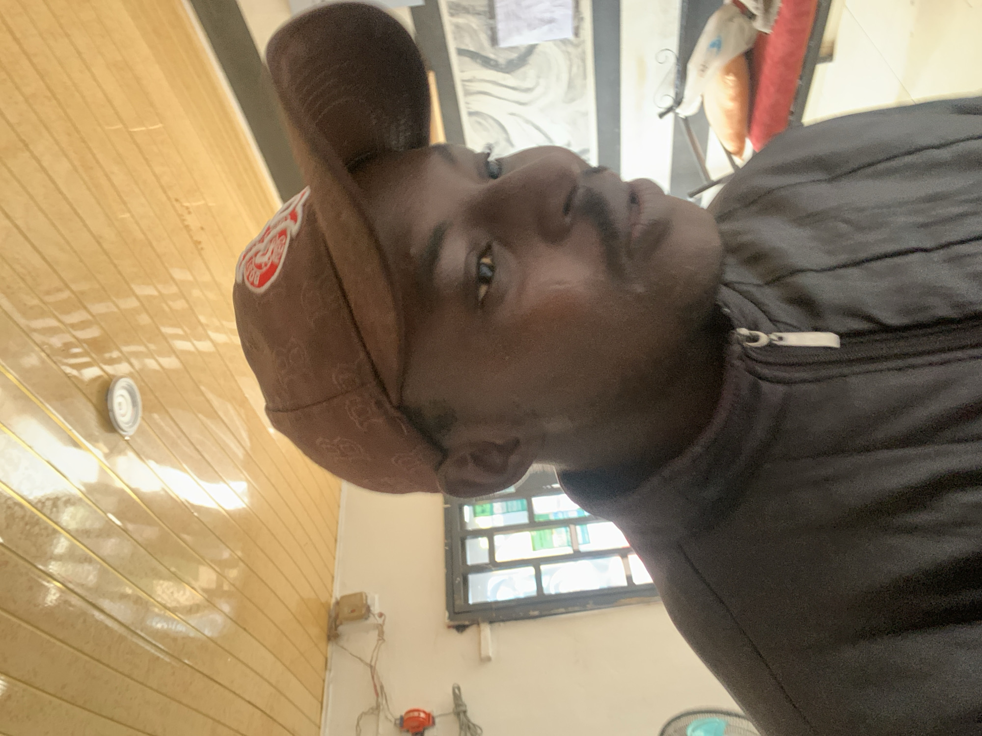 The image shows a close-up, candid shot of a person wearing a brown baseball cap and a dark jacket. The person's face is partially visible, with their eyes looking off to the right and their mouth slightly open. The background is blurred but appears to be an indoor setting with a wood-paneled ceiling and a window with metal bars. There are also some objects visible in the background, including what looks like a bed with pillows. There is no identifiable information or context that would place the scene specifically in Jos, Nigeria.