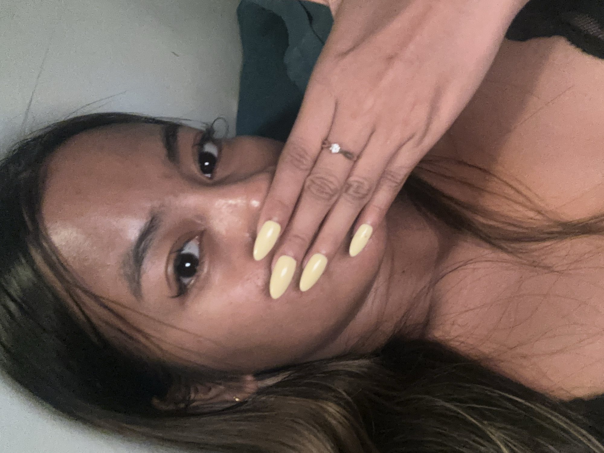 A close-up, selfie-style photograph shows the upper body of a young woman with medium-toned skin and dark hair. Her right hand, with pale yellow stiletto-shaped nails and a small, delicate ring with a clear stone, is covering her mouth. Her left eye is visible, with winged eyeliner and long lashes. The background is blurry and appears to be indoors, with a textured wall and a dark green fabric surface. The lighting is soft, creating a warm tone. The image was taken in Sukawati, Indonesia.
