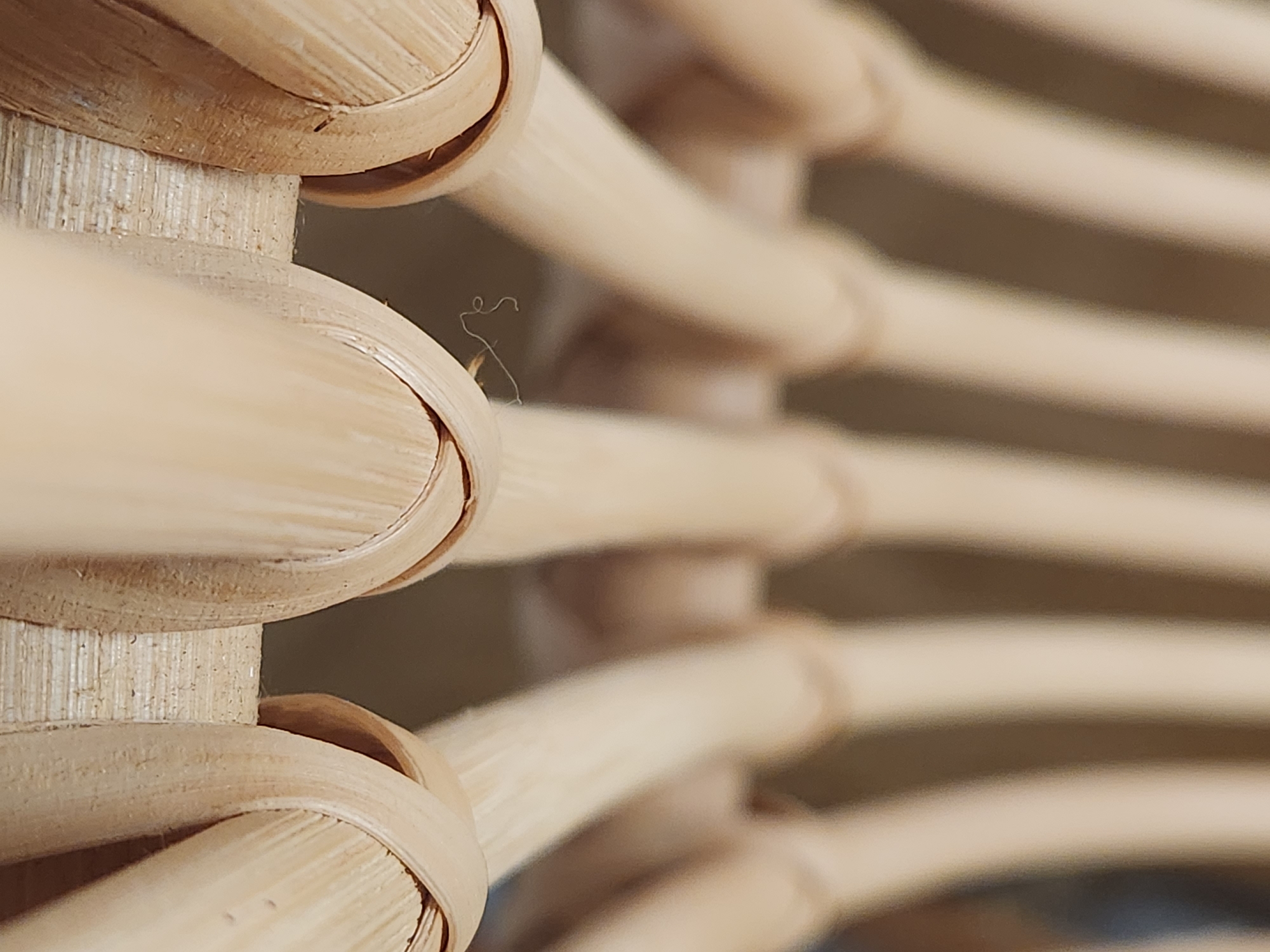 The image is a close-up, abstract shot of a woven material, likely bamboo or rattan. The subjects are the interwoven strands of the material, forming a textured surface. There is no discernible activity or event taking place; the focus is on the intricate pattern and natural textures.
Notable details include the light brown and beige tones of the material, with subtle variations in color and texture where the strands overlap and are woven together. The lighting suggests an indoor setting with soft, diffused light, casting gentle shadows that highlight the three-dimensional nature of the weave. A few wisps of fine, light-colored fibers are visible, adding to the organic feel. The background is out of focus, appearing as blurred bands of color that do not offer specific location cues. There is no visible text in the image. The image evokes a sense of natural craftsmanship and organic design.