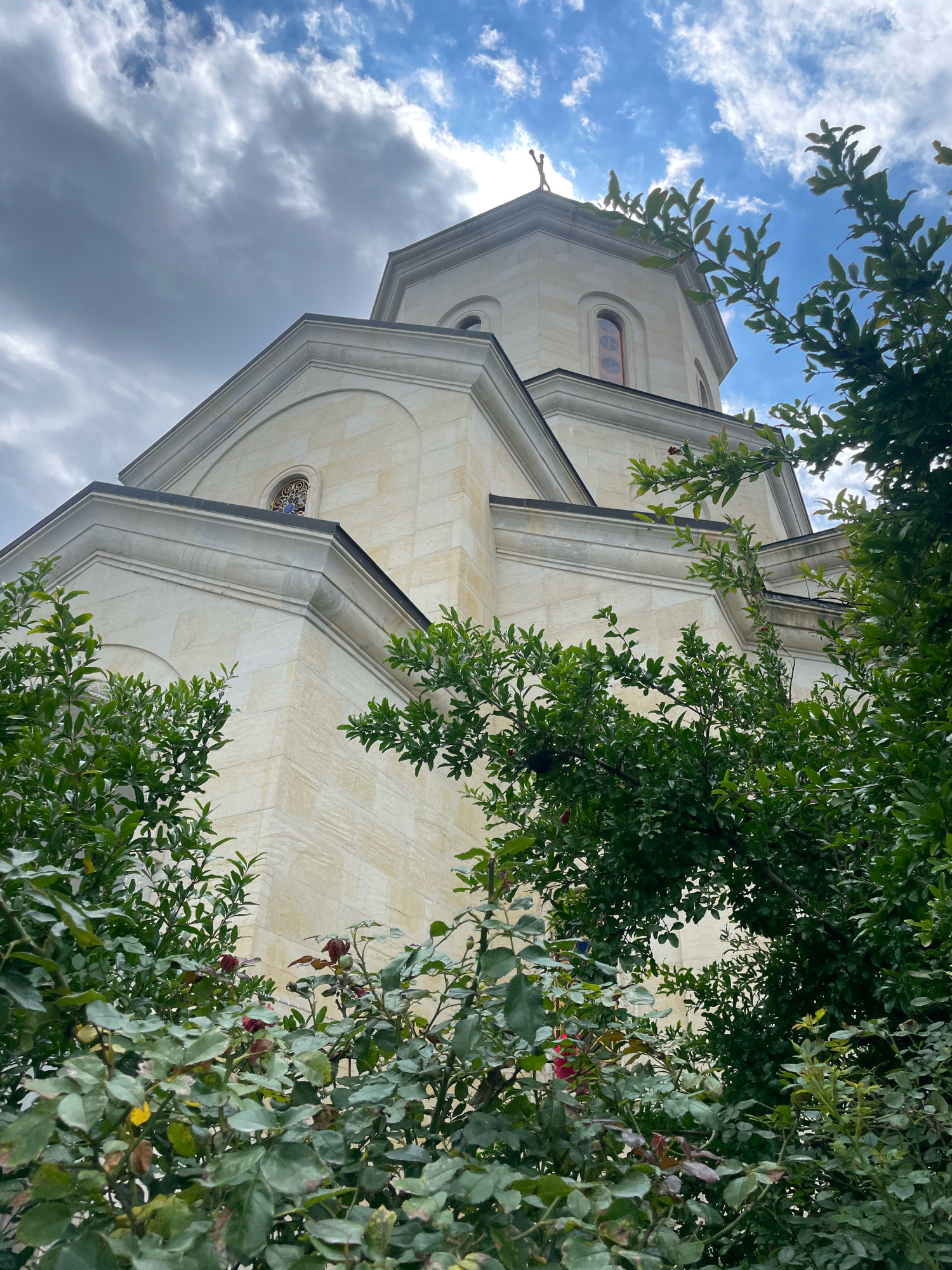 The photograph captures an upward-angle view of a cream-colored stone church structure, possibly a monastery or cathedral, set against a partly cloudy sky. Lush green foliage, appearing to be trees and bushes with some red flowers, frames the lower and right portions of the image, partially obscuring the building. The architecture features geometric lines, curved roof segments, and arched windows. A cross is visible at the apex of the main dome. The scene is devoid of any people. The image is a static, outdoor, architectural and natural landscape shot. The building's design suggests it is a religious edifice in Saguramo, Georgia.