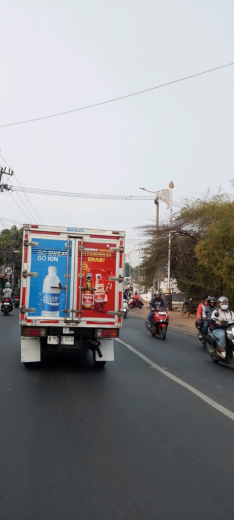 In Cileungsi, Indonesia, a white box truck with red trim drives on a paved road. The truck's rear doors display advertisements: the left door features a large image of a Pocari Sweat bottle with the text "GO SWEAT GO ION", and the right door shows two bottles of Vitamin C drink with cartoon characters and the text "VITAMIN C ENAKI!". The truck has a license plate reading "B 9310 SCD". Several motorcycles are visible on the road: one is ridden by a person wearing a helmet and a grey jacket in front of the truck on the left; another motorcycle driven by a person in a red jacket is to the right of the truck; and a group of three motorcycles with riders wearing helmets are further to the right, one with a rider in a grey jacket and another with a rider in a white jacket and helmet. The environment includes utility poles and wires overhead, and a tree line with foliage on the right side of the road. The sky is overcast.