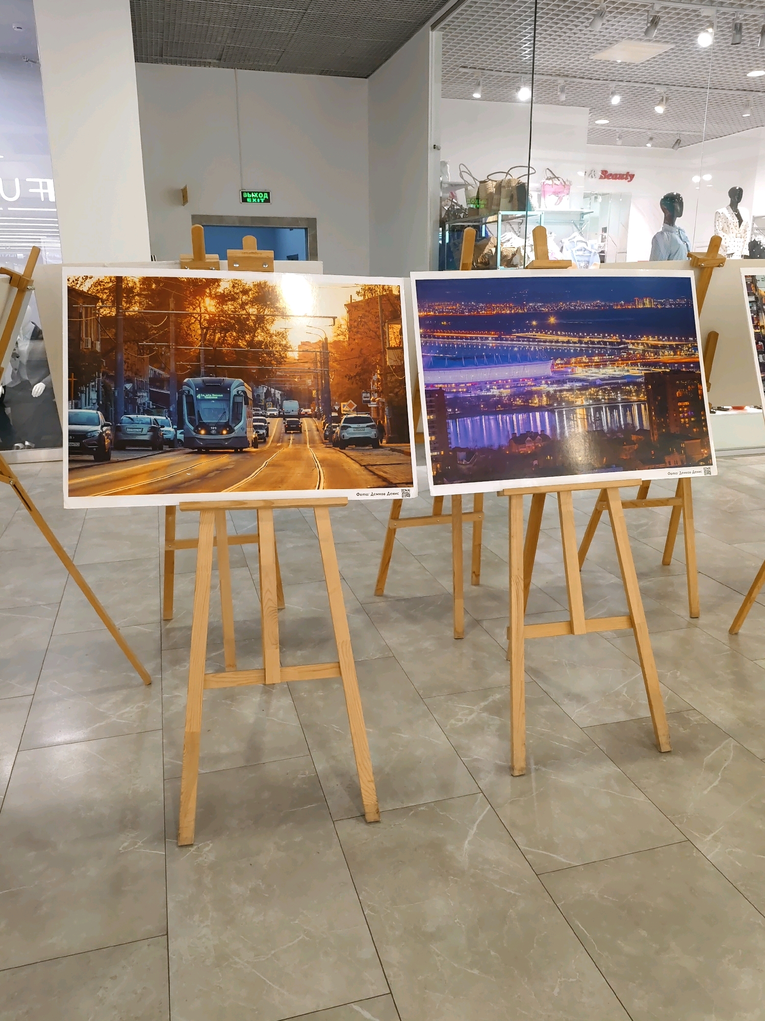 This image depicts an indoor exhibition of photographs mounted on wooden easels, likely situated within a commercial space in Rostov-on-Don, Russia. The photographs showcase urban scenes. One prominent photograph on the left displays a tram on a street bathed in warm, golden hour sunlight, with cars and buildings lining the road. Another photograph to the right captures a city skyline at night, illuminated by numerous lights reflecting on a body of water. In the background, behind the easels and through a glass storefront, a retail environment is visible, featuring mannequins, clothing racks, and signage, including what appears to be a "Seauty" store. Above an entryway in the background, a green illuminated "EXIT" sign is present. The flooring is tiled, resembling marble. No people are clearly identifiable within the exhibition space itself, though figures might be present in the background retail area.