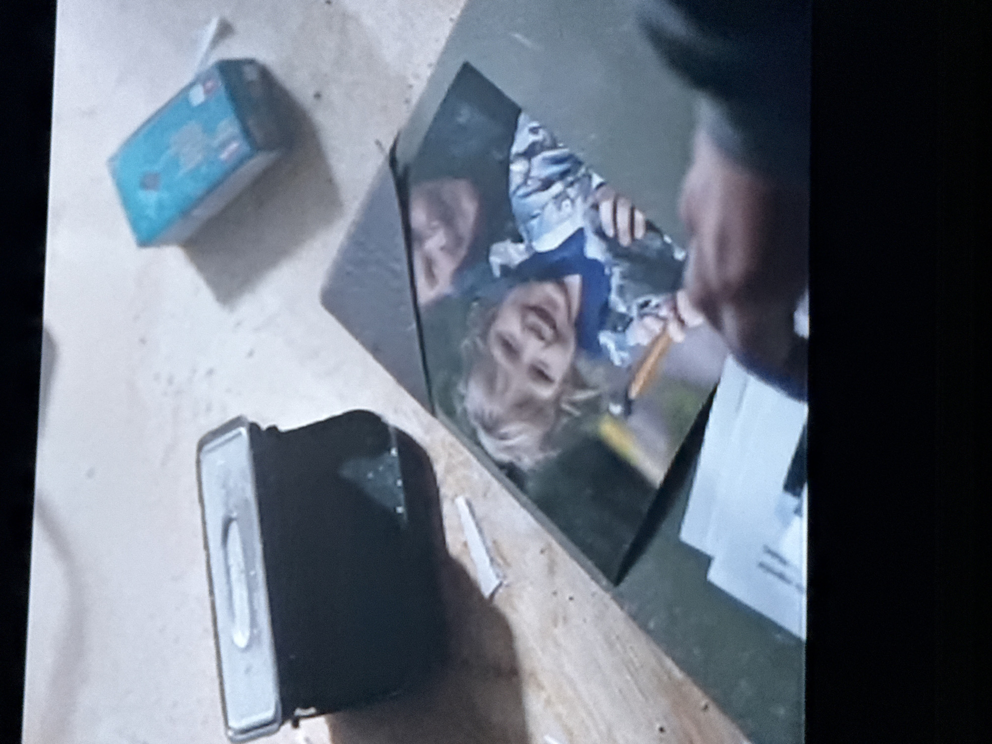 A cluttered scene includes a small blue box, a dark-colored rectangular object with a silver top, a photograph of a child with an adult, papers, and an open green folder. The location and city details are not determinable from the image.