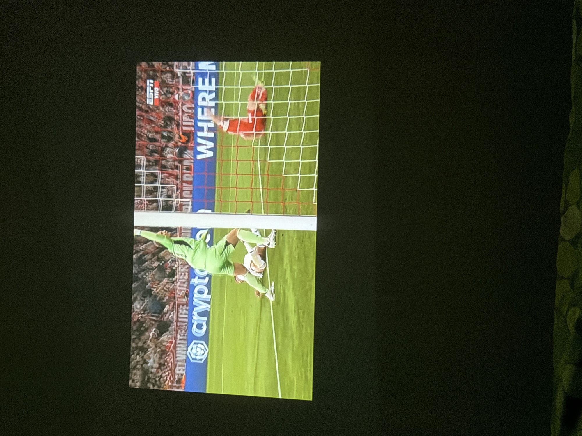 The image shows a soccer game being displayed on a screen. A player in red is in the goal being scored on. The opposing goalkeeper in green and white uniform is saving a shot. The advertisement on the side of the field is for "crypto.com". The broadcast is on ESPN. A sign states "WHERE." I do not have the information to determine if the game took place in Milan.