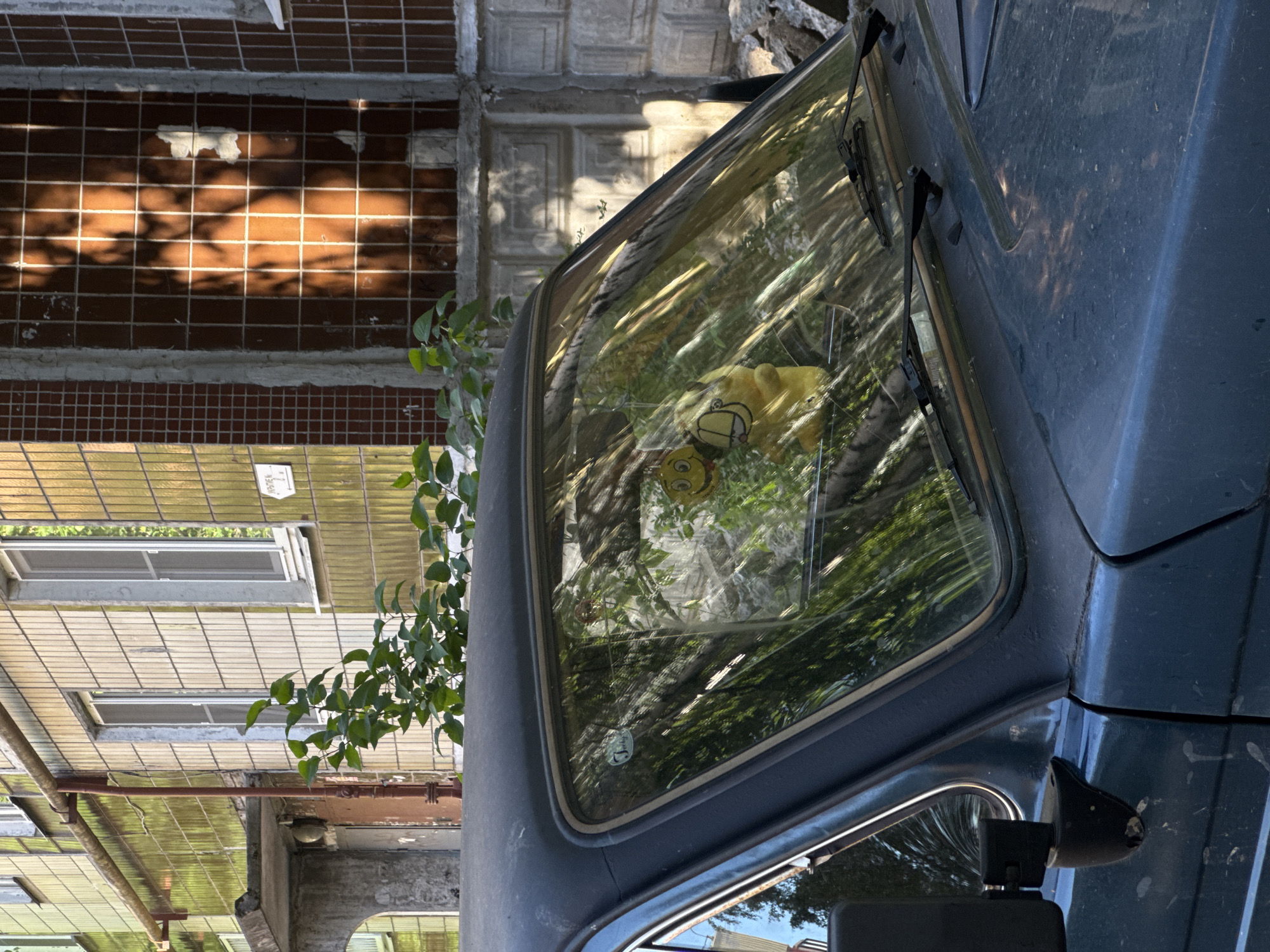 An outdoor, eye-level, static shot captures the front of a dark blue, dusty vehicle, likely a car, parked in an urban environment. The windshield, reflecting green foliage and parts of the background buildings, displays two yellow novelty items: a circular smiley face with wide eyes and a yellow Garfield plush toy wearing a small hat, both hanging inside. A circular white sticker is affixed to the upper left of the windshield. Two black windshield wipers rest horizontally at the base of the glass. The car's dark roof and a black side mirror on the left are also visible.
In the background, two multi-story buildings stand under direct, strong sunlight, casting distinct shadows. The building to the left features a facade of alternating light beige and greenish-yellow rectangular tiles, with several white-framed windows. A rusted metal pipe structure runs down its side, and a concrete canopy partially covers an entrance. A white sign with Cyrillic text "УКРЫТИЕ № 2" (Shelter No. 2) and an arrow is mounted on this building. The building to the right is covered in smaller, dark red-brown square tiles and features a large, rectangular opening covered by a metal grid, revealing a lighter interior with some peeling white material. Below the tiled section on the right, a concrete structure with decorative relief is visible. Green leafy foliage partially obscures the buildings between them and the vehicle. Some broken concrete and rubble are present on the ground to the far right. The overall scene suggests an urban setting in the city of Unknown, Unknown. No people are present.