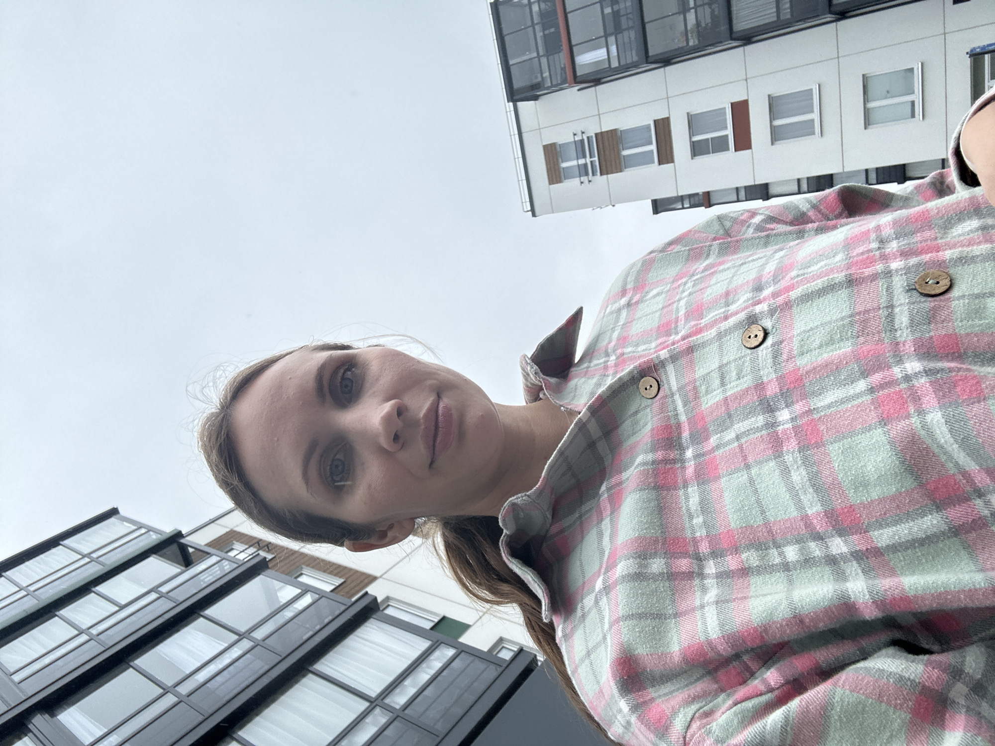 A low-angle, medium shot captures a woman with long brown hair pulled into a ponytail, wearing a pink and light green plaid shirt. She is looking at the camera with a neutral expression. Behind her, modern apartment buildings with dark-framed windows and balconies are visible against an overcast sky. The image appears to be taken outdoors, possibly in a residential area of Vsevolozhsk, Russia, given the architectural style of the buildings.