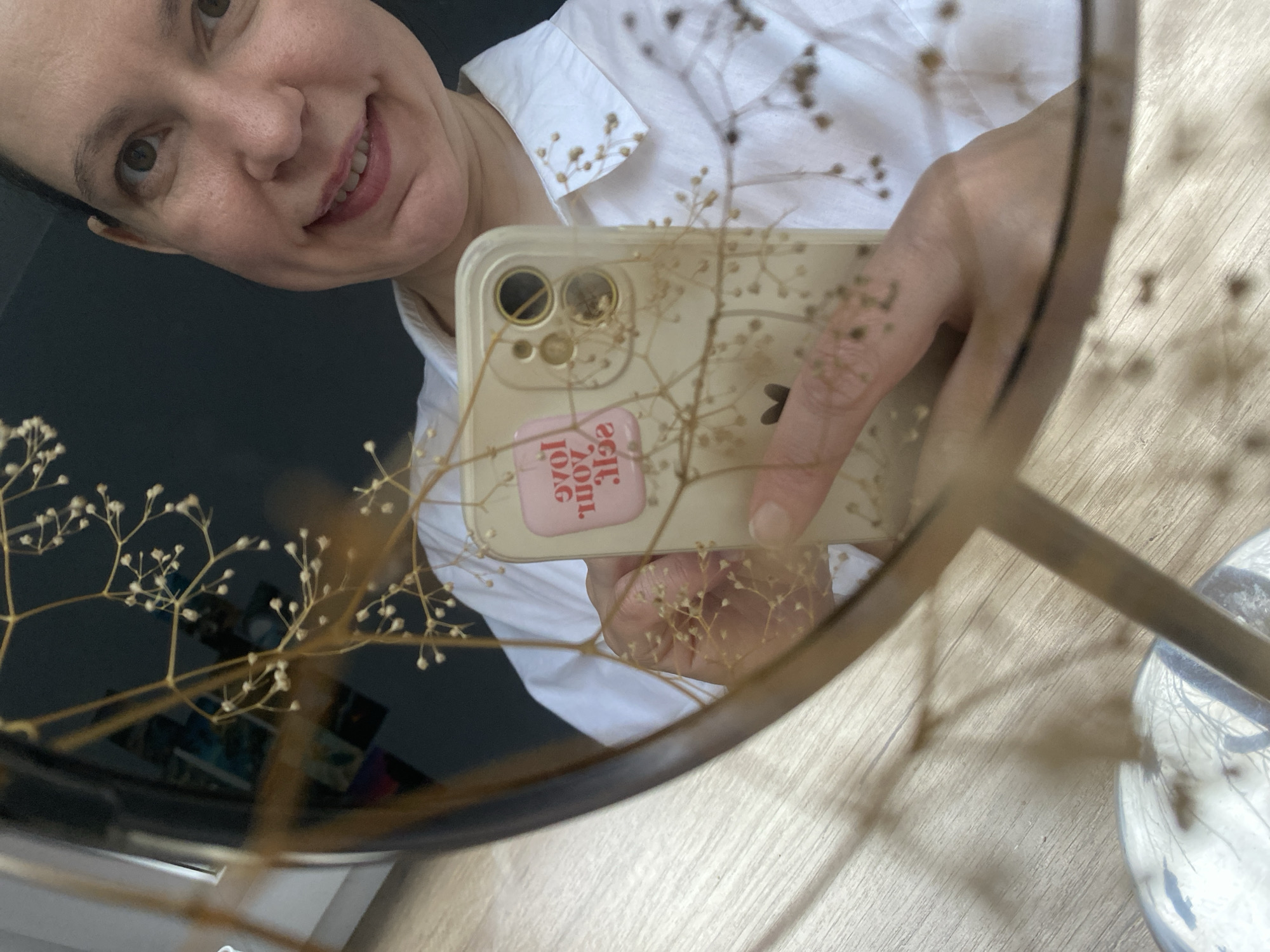 A woman with short, dark hair and a white collared shirt takes a selfie in a round mirror. She is holding a gold iPhone with a clear case and a sticker that reads "I love you so much." Dried baby's breath flowers are obscuring parts of the phone and mirror. The background of the reflection includes a dark surface and a wooden surface with a white object on it. The image was taken in the city of Chaikovsky, Russia.