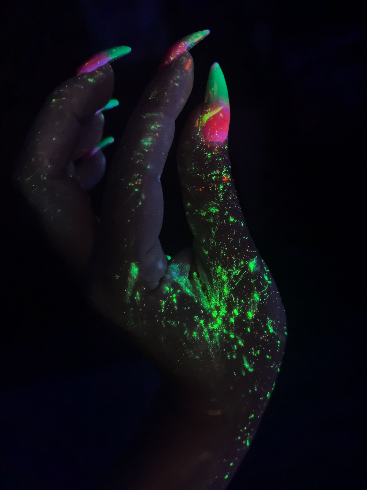 A human hand is prominently featured against a dark background, appearing as if illuminated by blacklight. The hand is covered in speckles and patterns of green and orange light, seemingly the result of fluorescent or phosphorescent material. The fingernails are long and painted with a two-tone effect, transitioning from a pinkish-red at the base to a vibrant green at the tips. The fingers are slightly curled, and the hand is angled upwards. The lighting suggests a nightlife or party environment, and the effect creates a surreal and artistic aesthetic. The image provides no information on the location within Noviy, Russia.