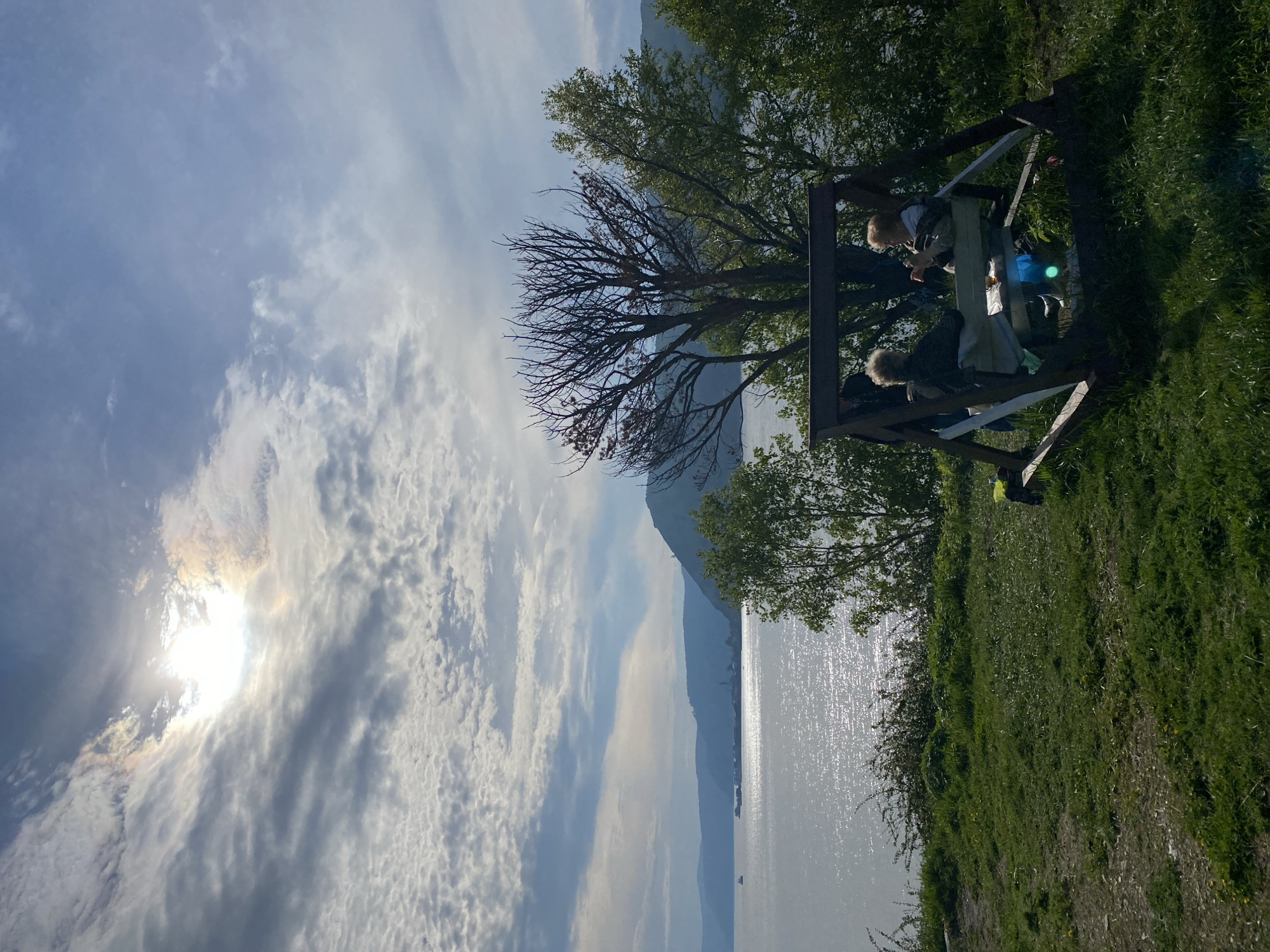 Two elderly individuals are seated on a wooden swing-bench facing the viewer. The swing is positioned on a grassy hill overlooking a wide body of water, possibly Lake Baikal. The water reflects the sunlight, creating a shimmering effect. In the background, rolling hills and mountains are visible, shrouded in a light mist. The sky is a mix of blue and white clouds, with the sun partially obscured by clouds, casting a radiant glow. Trees, some bare and others with green foliage, surround the swing and the edge of the hill. The overall scene suggests a tranquil and scenic outdoor setting, likely in or near Listvyanka, Russia, given the characteristic landscape of the region.