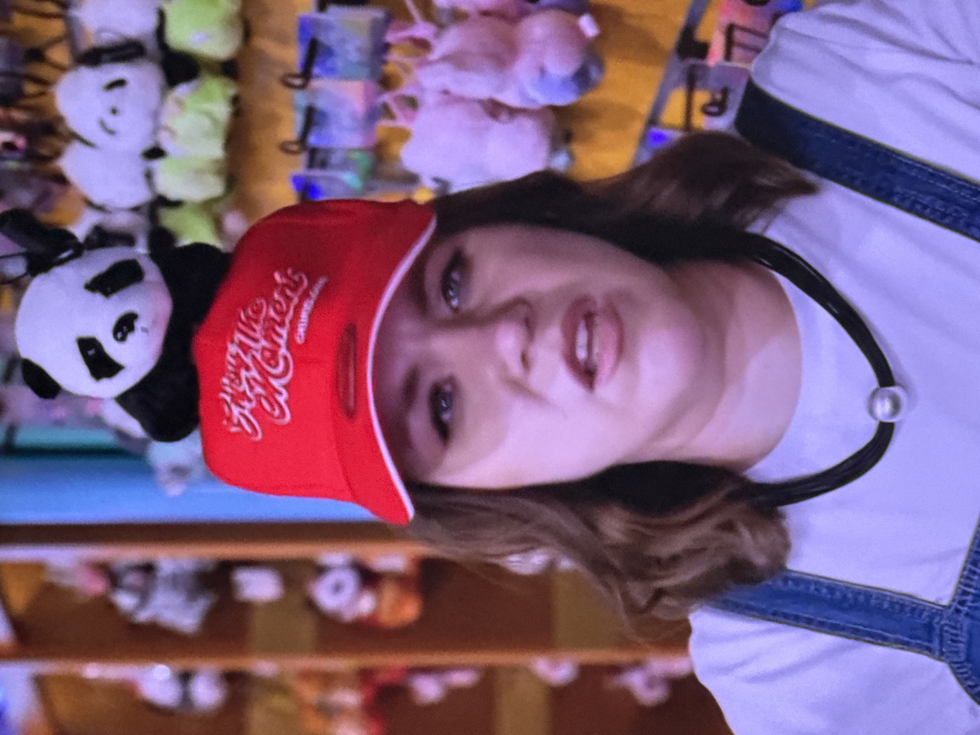 A young Caucasian woman with medium-length brown hair is shown from the chest up, wearing a red baseball cap with a panda plush toy perched on top. The hat has white lettering that appears to say "Seize the Moment" in a cursive font. She is also wearing blue overalls over a white t-shirt and a black choker necklace with a silver bead. Her facial expression is one of surprise or slight discomfort, with her mouth slightly open. In the background, there are shelves stocked with various plush toys, including more pandas and other animals, suggesting the image was taken in a store or arcade. The lighting is bright, and the overall atmosphere is casual and playful. There is no discernible location information within the image that would indicate Moscow, Russia.