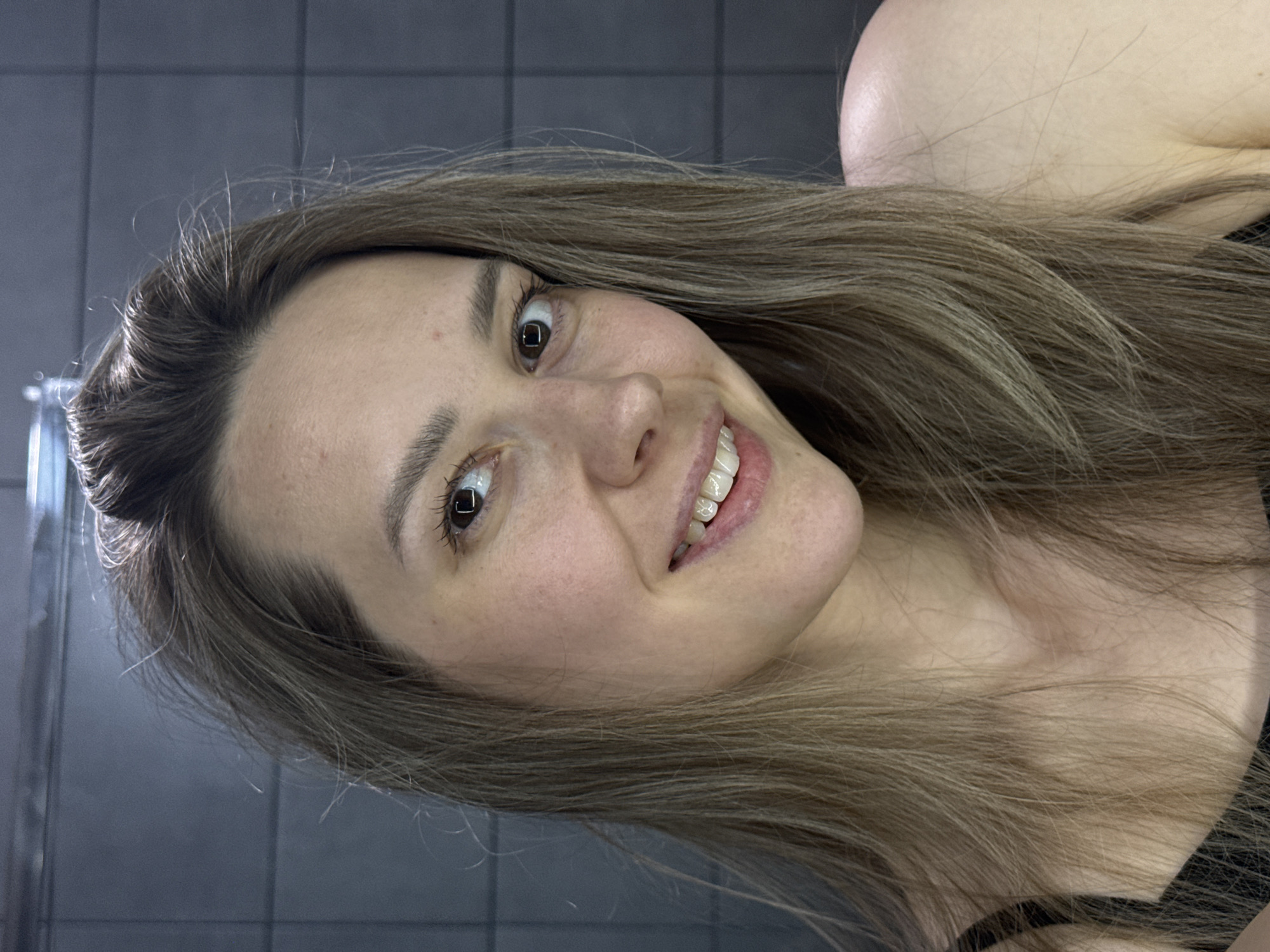 A close-up portrait shows a young woman with fair skin, light brown hair, and dark eyes, captured at a significant tilt. She is smiling broadly, revealing her upper teeth, and looks directly into the camera with a cheerful expression. Her long hair, styled with some loose strands, frames her face and falls over her shoulders. She is wearing a dark top, with a strap visible on her left shoulder. The background features dark grey square tiles, suggesting an indoor environment, possibly a bathroom or changing room in Chaikovsky, Russia. A reflective surface, possibly a mirror edge or shower glass, is partially visible on the far left. The lighting is artificial, illuminating her face. No visible text is present in the image.