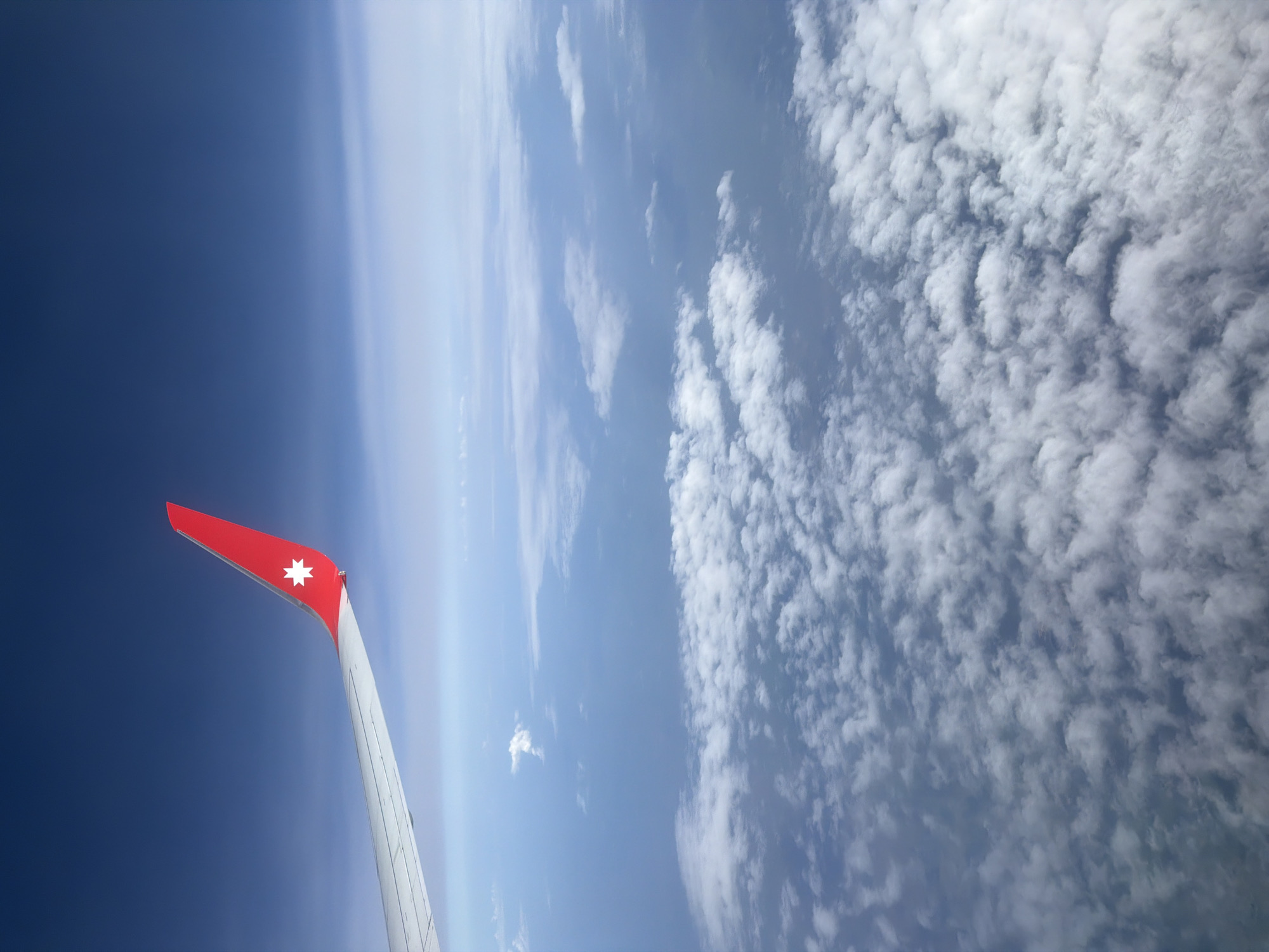 The image shows the wing of an airplane in flight, with a red tip and a white star on the tip. The background consists of a gradient of blue sky to the left, and fluffy white clouds filling the right portion of the frame. The plane's wing is angled upwards from the bottom left. No people are visible. There is no indication in the image to indicate the location as Domodedovo, Russia.
