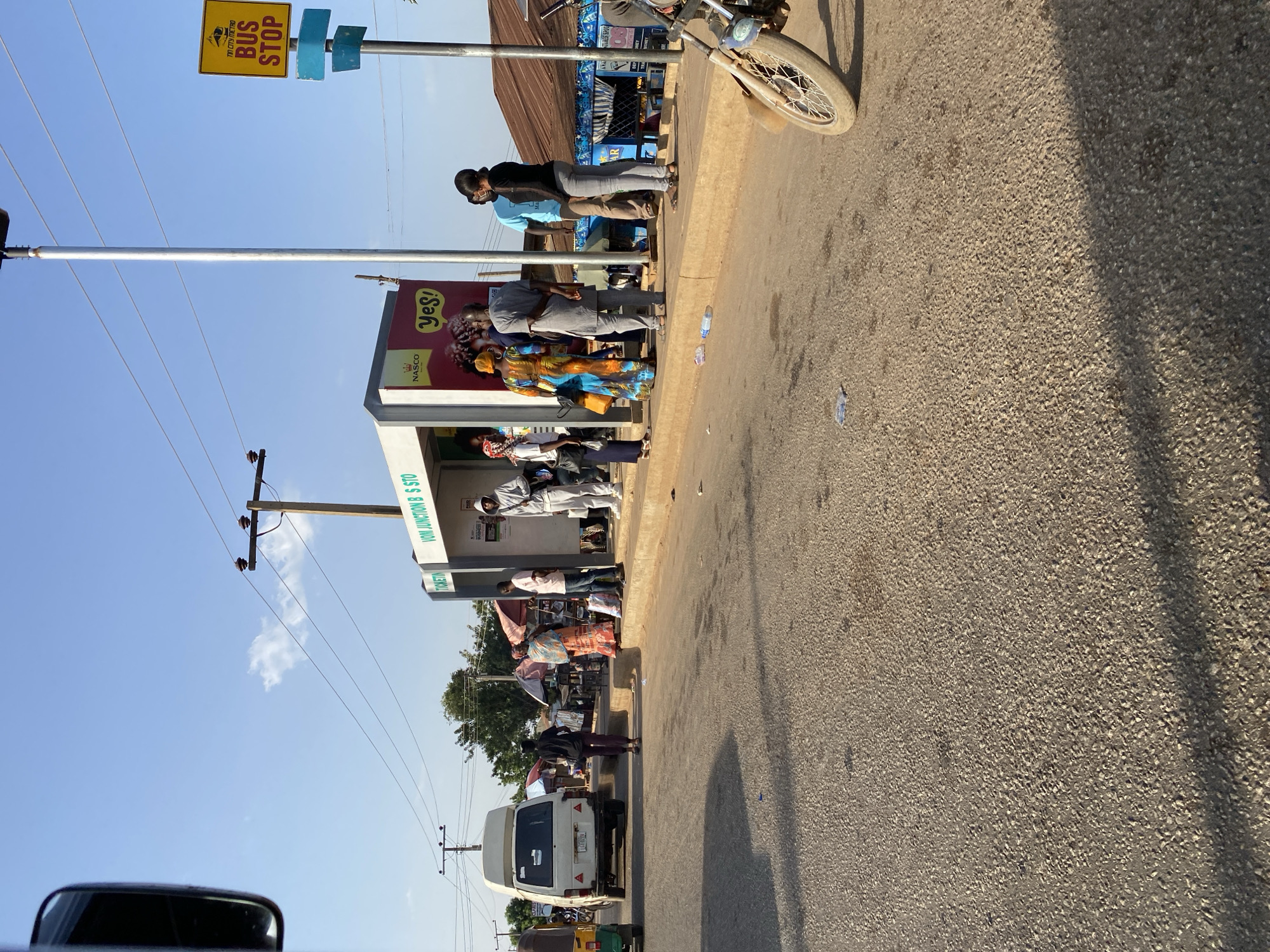 The image depicts a bus stop in Bukuru, Nigeria. The bus stop is marked by a yellow "BUS STOP" sign and a structure with advertisements, including one for "NASCO Yes!". People are waiting at the bus stop, some sitting and others standing. The road is paved and visible. There is a motorcycle parked near the bus stop. In the background, there are buildings, trees, and utility poles with power lines. A white van is visible further down the road. The sign on the bus shelter reads "VOM JUNCTION B S STO TICKETIN".