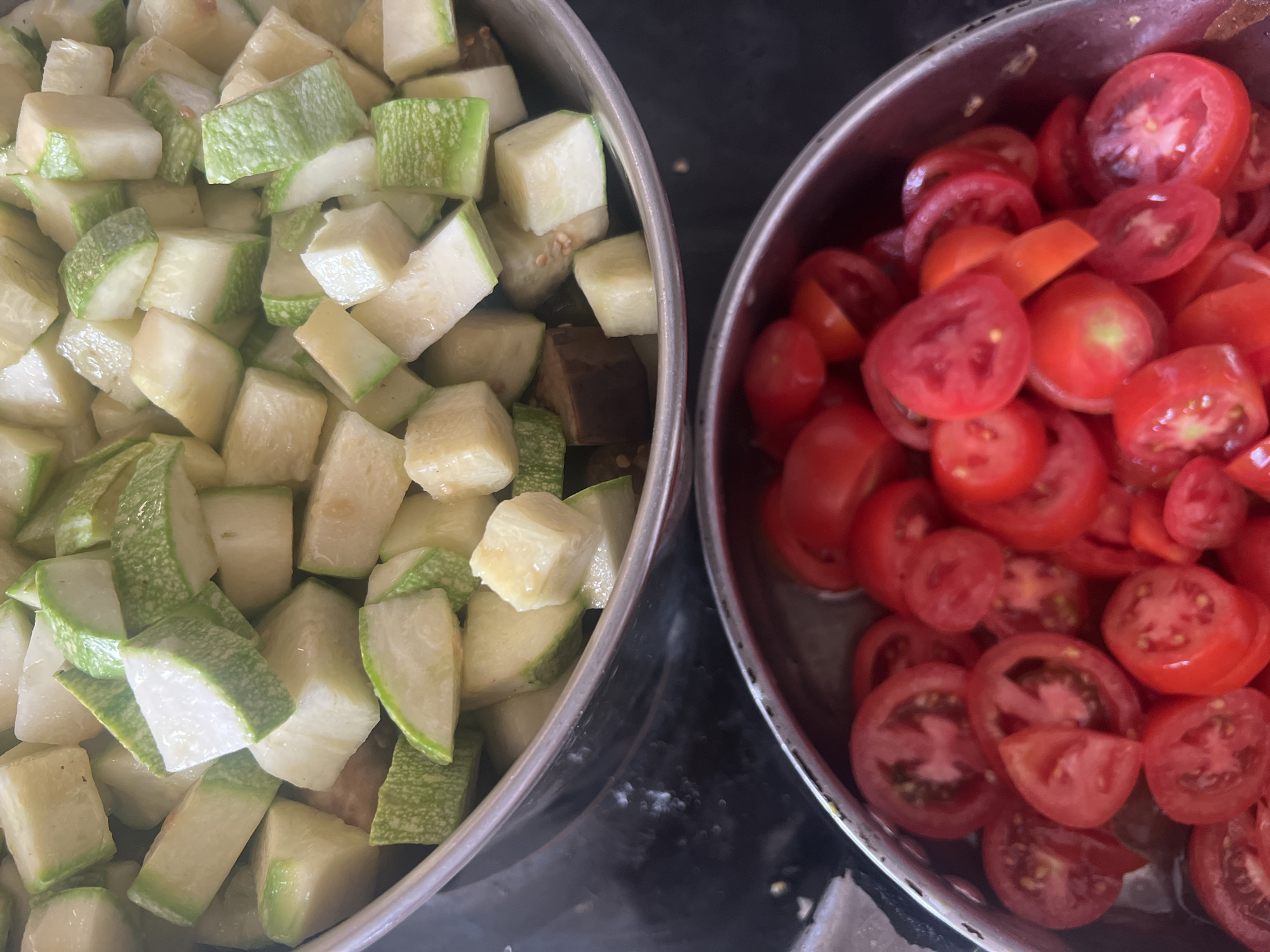 Two metal bowls are filled with chopped vegetables. The top bowl contains diced zucchini and eggplant. The bottom bowl is filled with sliced cherry tomatoes. The background is dark and out of focus. The scene is an overhead shot.