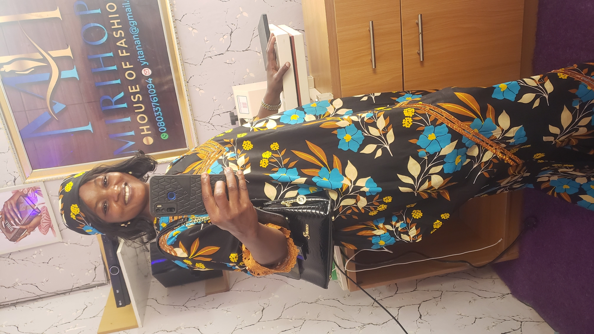 A woman is standing in what appears to be a business or retail space. She is holding a smartphone in her right hand and a black handbag in her left. She is smiling and looking at the phone. The woman is wearing a black dress with a floral pattern of blue and yellow flowers and brown leaves. She is also wearing a matching headscarf. Behind her is a framed sign with "MIRIHOP HOUSE OF FASHION" written on it, along with a phone number and email address. To her left, on a shelf, is a black electronic device. To her right, on a wooden dresser, is a white machine with a black plate on top. The walls have a light-colored, textured wallpaper with a delicate branch pattern. The floor is covered with a dark purple rug. The scene suggests a fashion boutique or a related business.