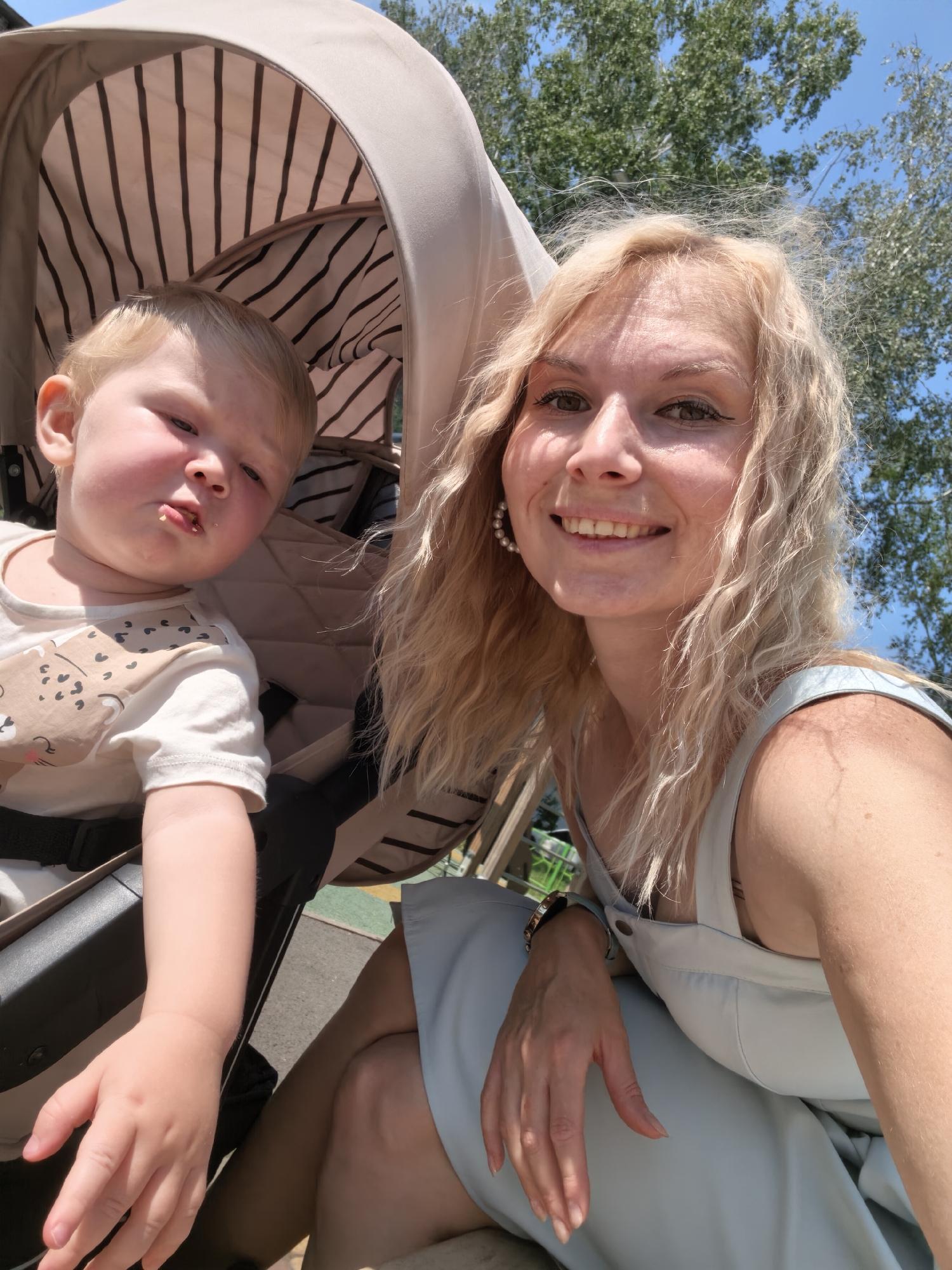 In a bright, outdoor setting, a woman with blonde wavy hair and a light blue dress is taking a selfie. She is smiling broadly, showing her teeth and looking towards the camera. She wears a large pearl earring in her left ear. To her left, a young child with blond hair sits in a stroller. The child is looking towards the camera with a slightly confused or surprised expression, and a small piece of food is visible on their lips. The stroller has a beige canopy with black and white stripes. The background consists of green trees and a clear blue sky, suggesting a park or a similar outdoor recreational area. The woman's left hand is resting on her knee, and she is wearing a watch on her left wrist. There is a partial view of the stroller's frame and the child's arm. The overall scene depicts a casual moment between a mother and child on a sunny day.