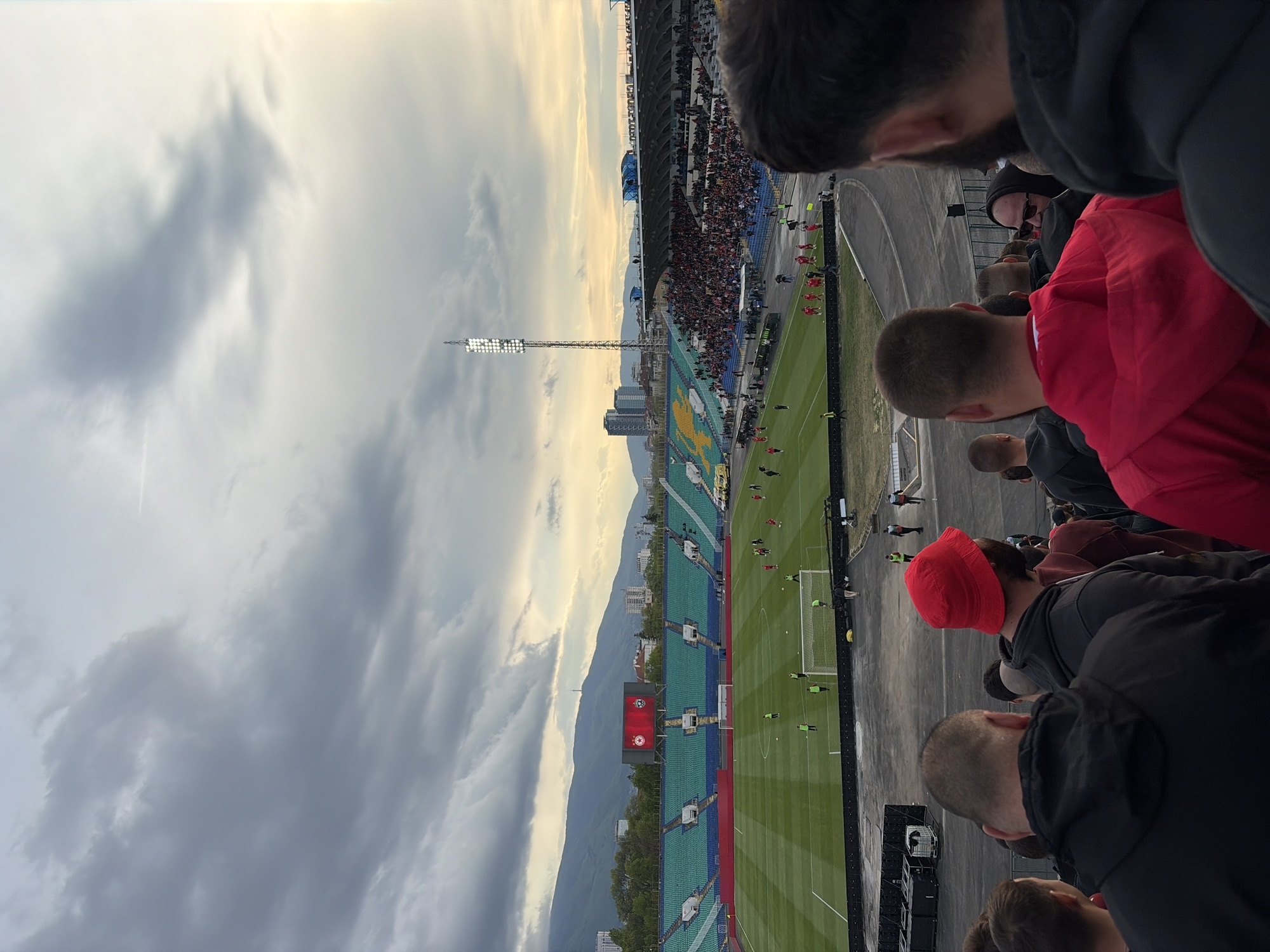 The image captures a wide view of a football stadium during a match, likely in the late afternoon or early evening. The perspective is from the upper stands, looking down towards the pitch and across to the opposing stands.
In the foreground, the backs of several spectators' heads are visible, most wearing dark clothing like hoodies. One person wears a bright red jacket, and another has a red beanie, indicating the viewer is among the crowd.
The midground features the vibrant green football pitch, where a match is in progress, with players scattered across the field. One goal is clearly visible on the right. The stadium stands are filled with numerous spectators, particularly on the opposite side. The seating appears to be primarily green and blue. A large scoreboard with a red background and a white emblem (possibly a team logo) is prominent on the far side of the pitch, below which faint white text is visible but illegible. Tall floodlight towers stand on either side of the field, with their lights illuminated, suggesting it might be getting dark or the sky is overcast.
In the background, a distinct mountain range stretches across the horizon under a dramatic, partly cloudy sky. The sun, casting a warm yellow glow, is breaking through the clouds near the horizon, contrasting with the darker, heavier clouds above. Some city buildings are visible between the stadium and the mountains. A flag, which appears to be the Bulgarian flag (white, green, red horizontal stripes), is visible on a pole near the scoreboard.
The scene depicts an active sporting event with a considerable audience, set against a scenic backdrop of mountains and a dynamic sky in what is identified as Sofia, Bulgaria.