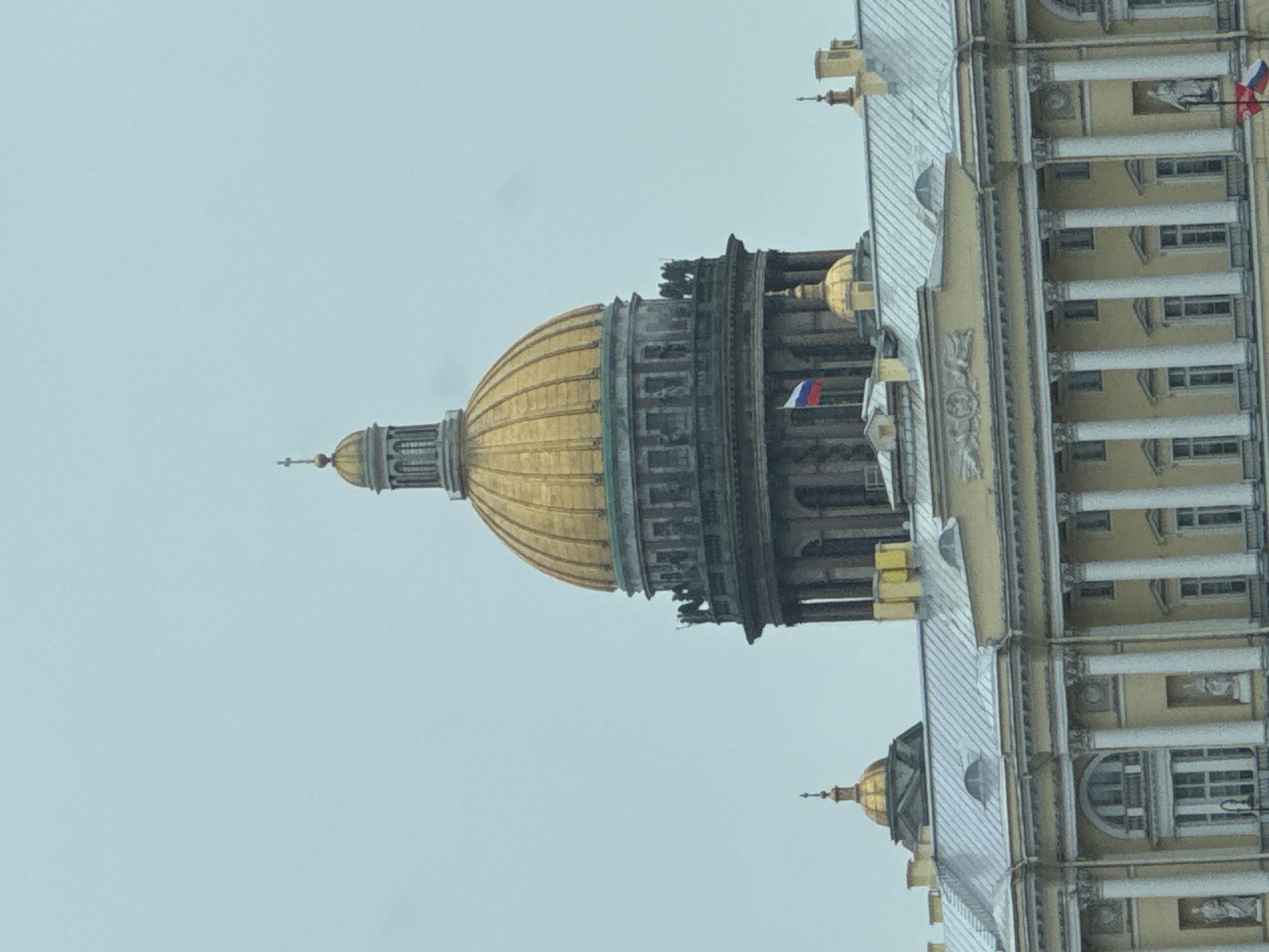 The image presents a grand architectural scene, dominated by the magnificent golden dome of a monumental building, strongly resembling St. Isaac's Cathedral in Saint Petersburg, Russia, as indicated by the location context. The large, gilded dome, complete with its upper lantern and spire, rises prominently against a light, overcast sky, showcasing its intricate structural details and the weathered patina on its lower drum.
To the right of the main dome, a significant portion of an elaborate building facade is visible. This section features a pale, likely beige or light yellow, exterior adorned with classical architectural elements such as towering white columns, decorative pilasters, cornices, and framed windows. The building's roof is a striking silver-grey, suggesting a metallic construction. A smaller, yet similarly styled, golden dome with a spire can be observed further to the right, adding to the building's stately appearance.
Several Russian flags, with their distinctive white, blue, and red horizontal stripes, are affixed to various parts of the structure, notably one near the main dome's base and another further along the building's facade, reinforcing the geographical location.
The overall lighting is soft and diffused, characteristic of an overcast day, with a uniform light blue-grey sky serving as the backdrop. There are no people visible, and no specific activities or events are taking place, creating a serene and timeless architectural study. The scene conveys an impression of historical grandeur and imposing beauty. No visible text is present in the image.