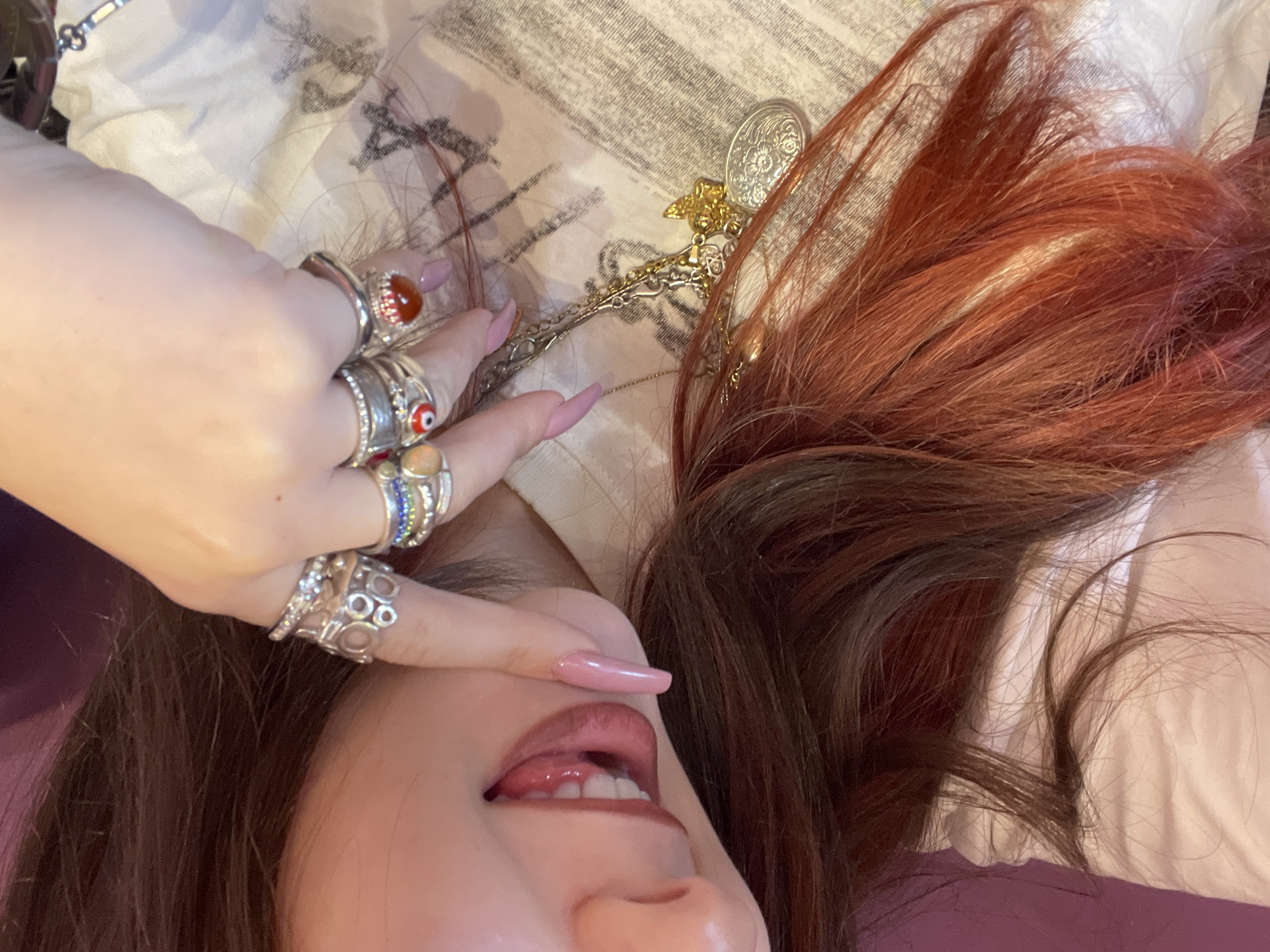 A close-up shot shows a person lying on a surface covered with a white and grey patterned fabric. The person's face is partially visible, with the mouth open and a tongue protruding slightly. One hand, adorned with multiple rings, is positioned near the face, obscuring part of the forehead and eye. The hair is a mix of brown and reddish hues. A silver and gold decorative chain with various pendants lies near the hair. It is not possible to determine the location as Raanana, Israel from the image.