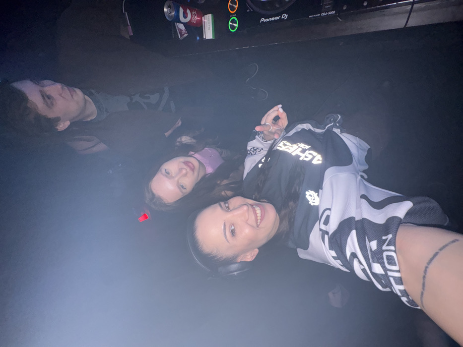 Three people pose in a dimly lit room. A man with dark hair is in the top left corner, looking towards the camera. Next to him is a woman with long brown hair, wearing a pink top. In the foreground, another woman smiles, wearing a black and white patterned jersey and headphones around her neck. A can of Pepsi and a Pioneer DJ CDJ-3000 player are visible in the background. A faint red light and smoky haze fill the room.